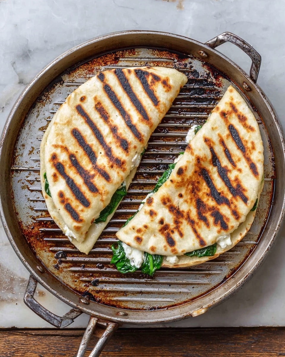 Two folded flatbreads with dark grill marks on top sit on a round metal grill pan with handles. Each flatbread is a light golden-brown color with a slightly crisp texture and has green spinach leaves and white melted cheese peeking out from the open edges. The grill pan shows oil stains and charred spots, adding a rustic look, and rests on a white marbled surface. photo taken with an iphone --ar 4:5 --v 7