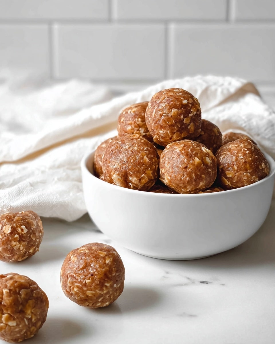 A white bowl filled with about ten round brown oat balls, showing a rough texture with visible oat pieces. Several more oat balls are scattered on a white marbled surface around the bowl. A white cloth is partially seen behind the bowl and a tiled white wall forms the background. The balls look dense and slightly shiny from natural ingredients. Photo taken with an iphone --ar 4:5 --v 7