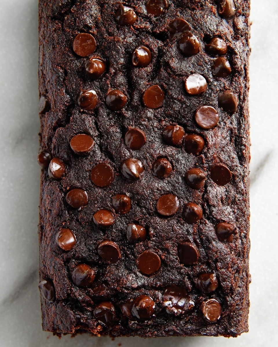 The image shows one thick, dark brown chocolate cake loaf with many shiny chocolate chips scattered unevenly on the top surface. The cake has a rough, moist texture with cracks and crevices giving it a rustic look. It sits on a white marbled textured surface with no other objects or decorations around. The chocolate chips vary in size and appear glossy, contrasting against the matte, dense cake base. photo taken with an iphone --ar 4:5 --v 7