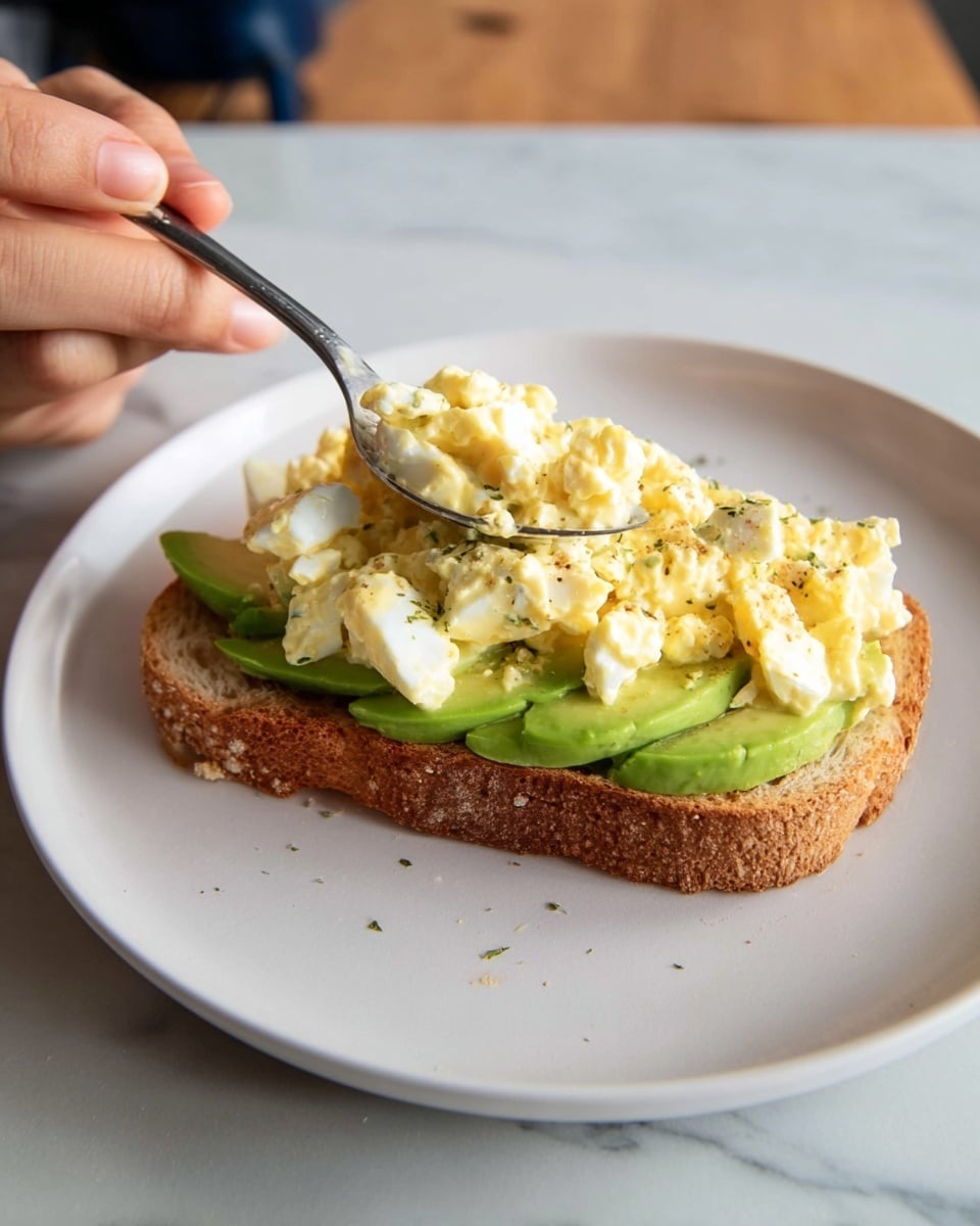 A slice of toasted bread sits on a white plate over a white marbled surface. On the bread, there is a layer of sliced avocado with its smooth green texture visible along the edges. On top of the avocado, there is a layer of creamy egg salad mixed with small chunks of egg white and yolk, displaying pale yellow and white colors with a soft, slightly chunky texture. A woman's hand is holding a spoon, spreading the egg salad evenly across the avocado layer. Photo taken with an iphone --ar 4:5 --v 7