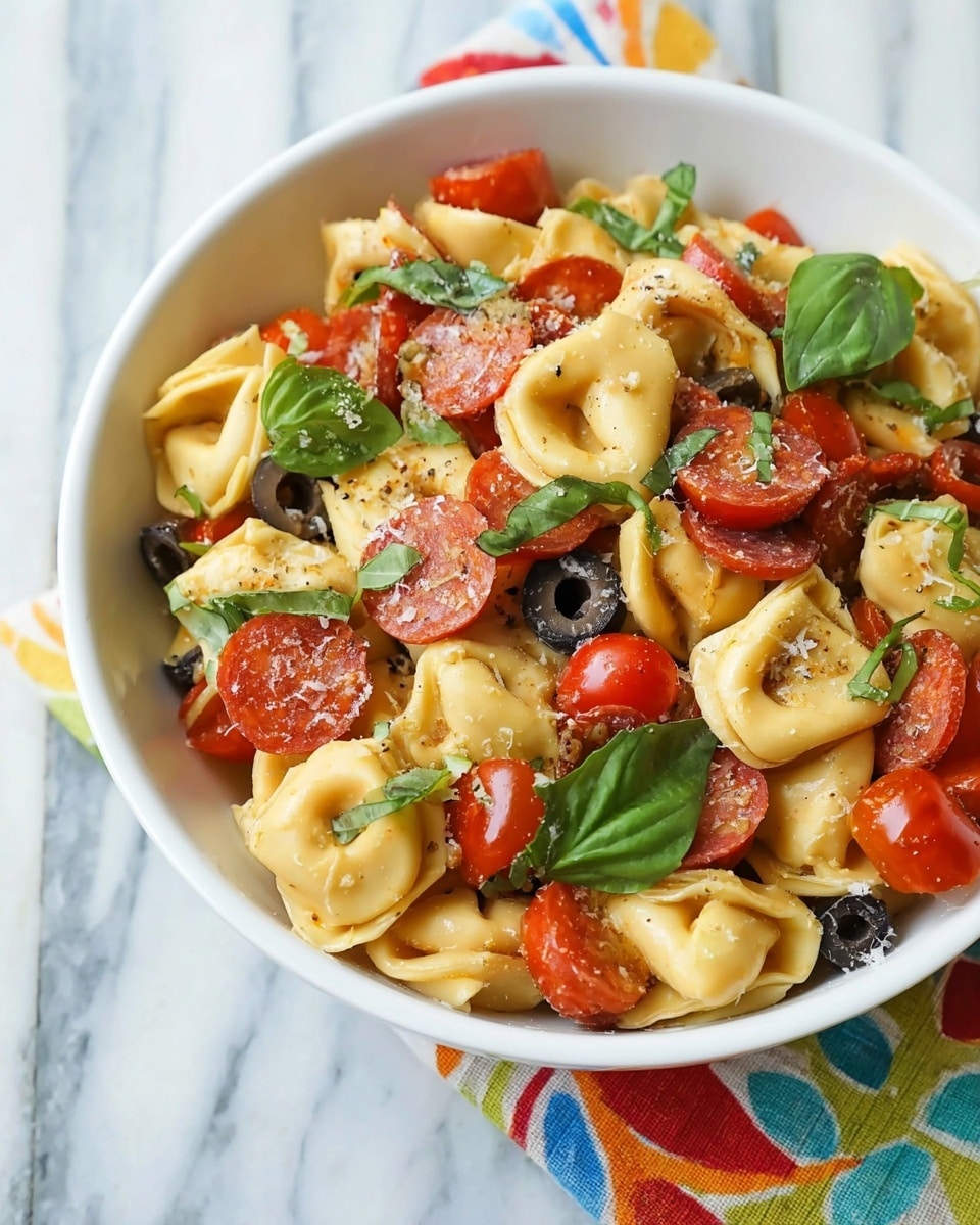 A white bowl is filled with tortellini pasta mixed with sliced black olives, halved bright red cherry tomatoes, and small round slices of orange pepperoni evenly spread throughout. Fresh green basil leaves are scattered on top, adding a touch of vibrant color. The pasta is coated lightly with a glossy sauce and sprinkled with bits of grated cheese, giving the dish a textured look. The bowl sits on a white marbled surface with a colorful cloth napkin partly visible under the bowl. Photo taken with an iphone --ar 4:5 --v 7