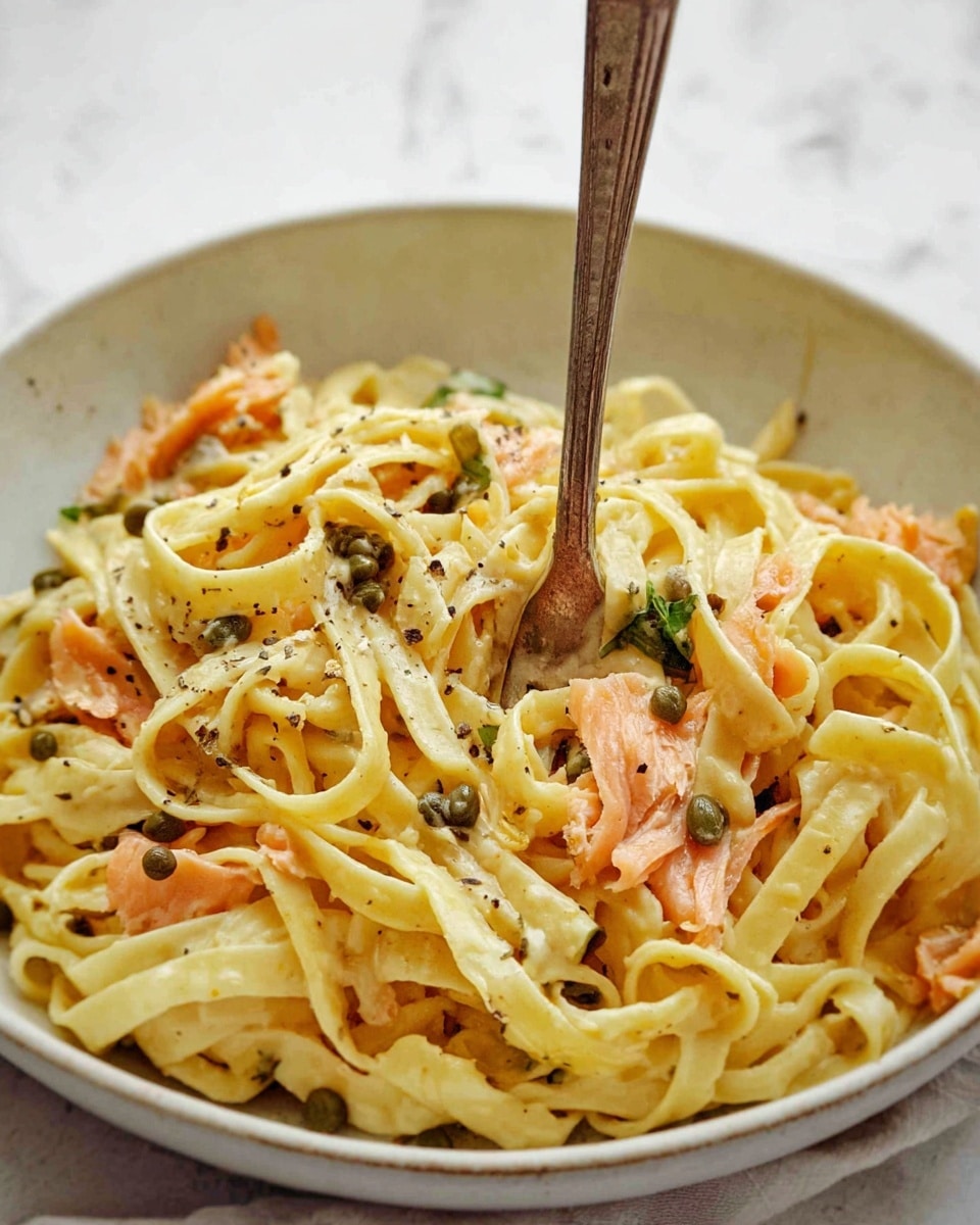The image shows a pan filled with creamy fettuccine pasta as the main base layer, pale yellow and coated in a smooth sauce. Scattered on top are pink-orange pieces of smoked salmon, adding vibrant color and a soft texture. Small dark green capers are spread evenly across the dish, giving a pop of color and contrast. Shredded white cheese is sprinkled lightly over the pasta and salmon, with cracked black pepper speckled all over. Fresh bright green basil leaves sit on the top, adding a fresh and lively touch. A silver spoon with an ornate handle rests in the pan on top of the pasta. The pan sits on a white marbled surface. photo taken with an iphone --ar 4:5 --v 7