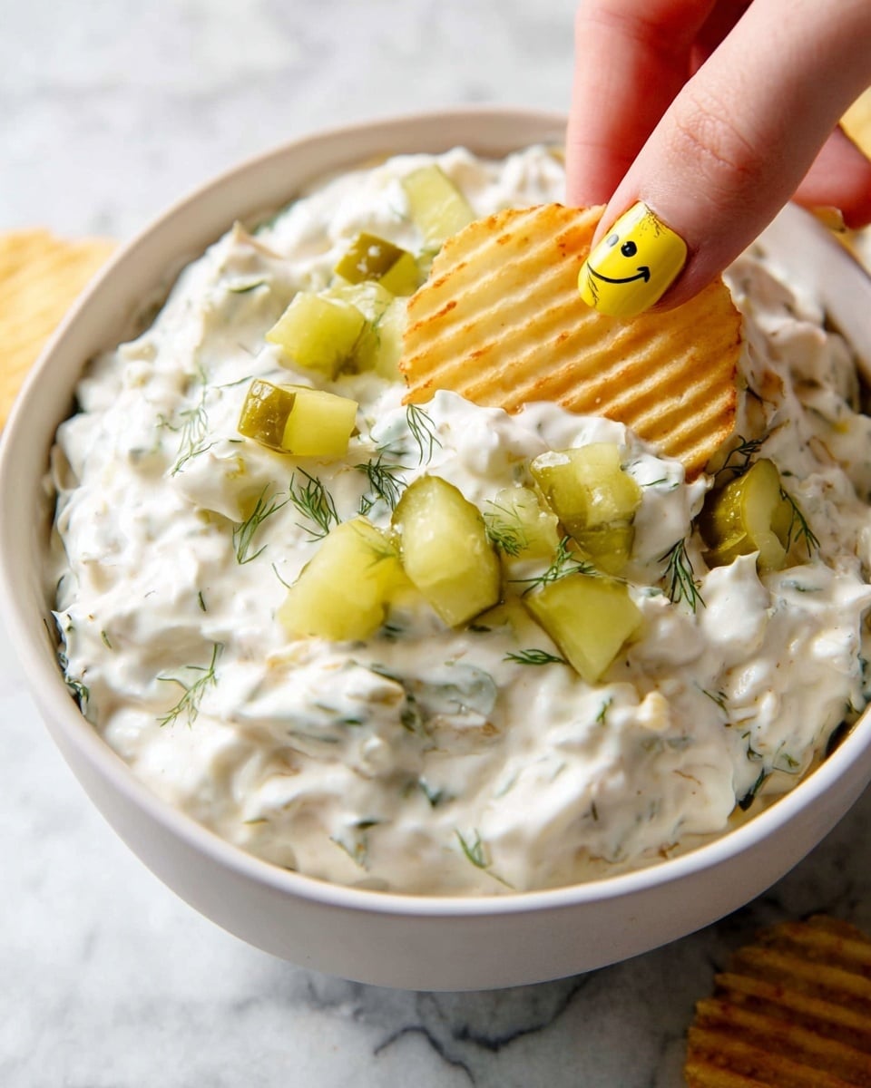 A close-up of a white bowl filled with thick, creamy white dip that has small green herb pieces mixed throughout. On top of the dip are scattered small cubes of bright green pickles and some darker green dill flecks. A woman's hand with a yellow nail showing a black smiley face design is dipping a ridged, golden-yellow potato chip into the creamy dip. The background shows a white marbled texture. photo taken with an iphone --ar 4:5 --v 7