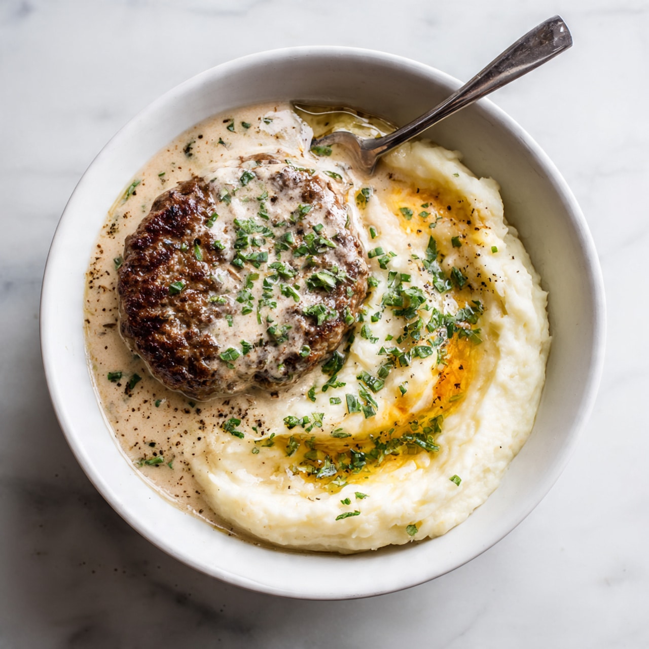A simple white bowl holds two main layers: a browned, grilled meat patty on the left, covered partially with a creamy, light beige sauce with specks of herbs, and on the right, a mound of smooth mashed potatoes topped with melted butter pooling in the center and sprinkled with fresh green herb bits and black pepper dots. A silver fork rests inside the bowl, leaning on the right edge. The background is a white marbled texture. Photo taken with an iphone --ar 4:5 --v 7