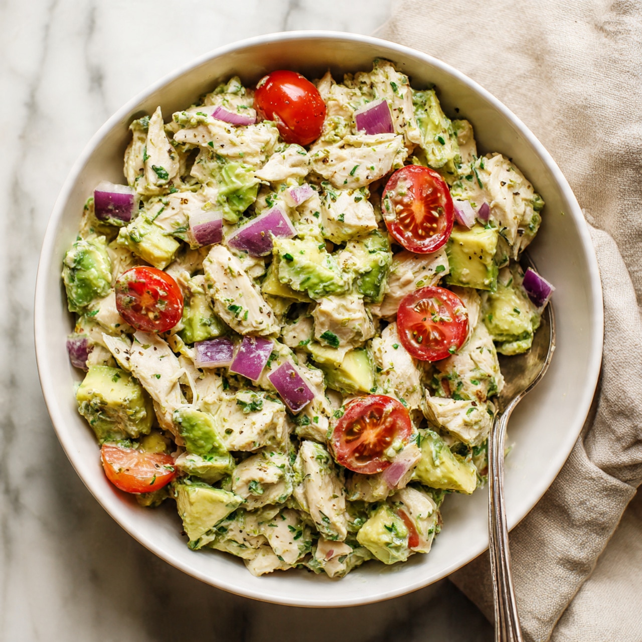A white bowl filled with a chicken salad showing several layers and colors, starting with chunks of white chicken mixed with light green avocado pieces. Small, bright red cherry tomato halves and small purple onion pieces are spread evenly throughout the salad. The mixture looks creamy with a hint of green from herbs or dressing blending all the ingredients. To the side inside the bowl is a silver fork resting on the edge. The bowl is on a white marbled surface with a light beige cloth partially visible next to it. photo taken with an iphone --ar 4:5 --v 7