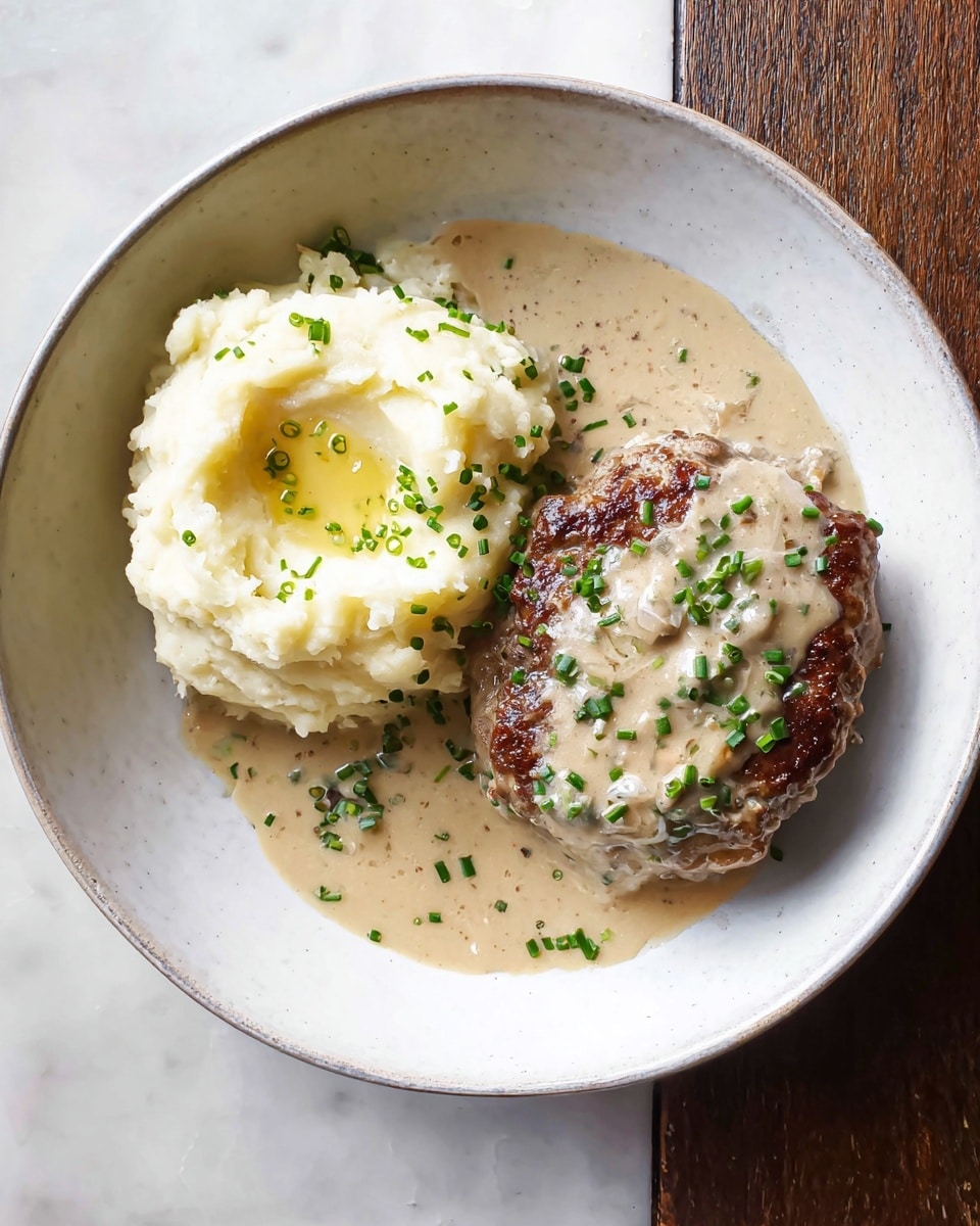 A white bowl holds two main parts: on the left, a mound of creamy mashed potatoes with a small pool of melted butter in the center, sprinkled with small green chives; to the right, a thick browned patty covered partially by a light beige creamy sauce with visible bits and more green chives scattered on top and around the sides. The plate sits on a white marbled surface. photo taken with an iphone --ar 4:5 --v 7