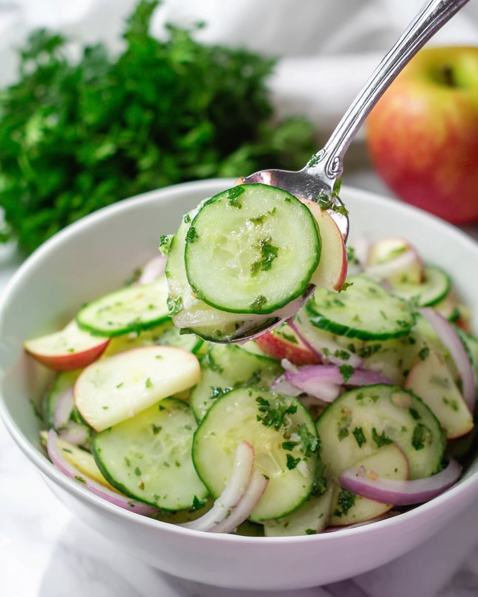 A white bowl filled with a fresh cucumber salad sitting on a white marbled surface. The salad has thin green cucumber slices layered on top, mixed with small red apple pieces, thin light purple onion slices, and bits of green parsley spread evenly throughout. A silver spoon is lifting a portion of the salad, showing a clear cucumber slice with apple pieces, onion slivers, and green herbs on it. In the background, blurred green leafy herbs and a whole apple are visible. photo taken with an iphone --ar 4:5 --v 7