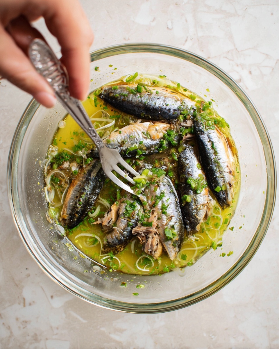 A clear glass bowl sits on a white marbled surface, filled with five pieces of small fish layered evenly around the edges. The fish have dark shiny skin with some silver and light brown patches. In the center of the bowl, there is a thick yellowish-green sauce mixed with small green chopped herbs and thin slices of green onion scattered around the fish. A silver fork held by a woman's hand is pressing down into the ingredients, mixing them together gently. The scene shows a close-up top view, capturing the texture of the fish skin, the sauce’s slight bubbles, and the freshness of the chopped herbs photo taken with an iphone --ar 4:5 --v 7