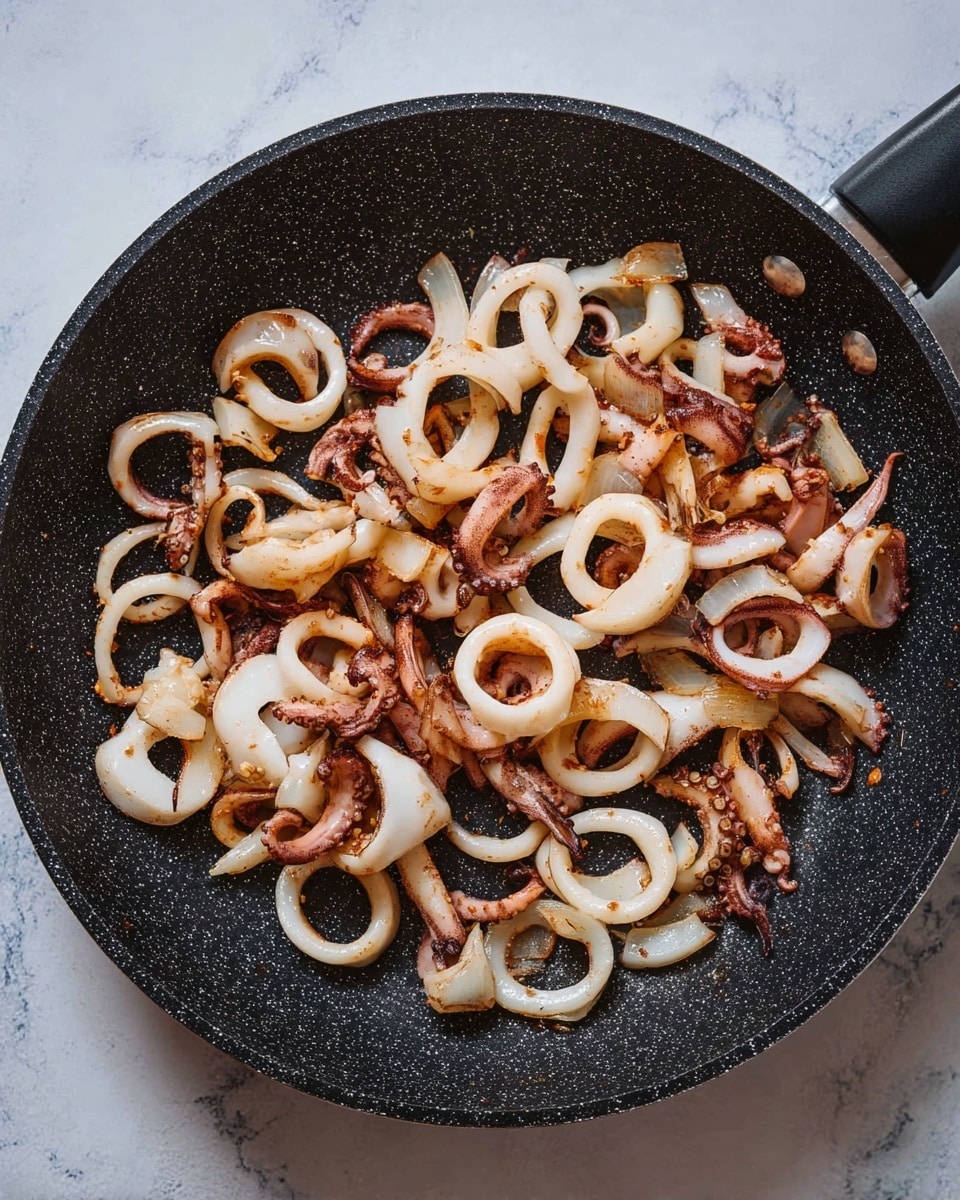 A black speckled frying pan filled with cooked squid rings and tentacles spread evenly inside. The squid pieces are white with light brown grill marks and some slightly darker browned spots, showing a mix of round rings and curled tentacles across the pan. The pan is placed on a surface with a white marbled texture. Photo taken with an iphone --ar 4:5 --v 7