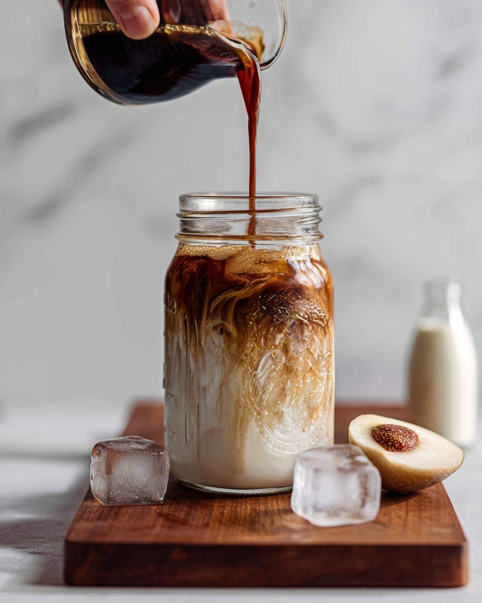 A tall clear glass jar filled with three visible layers: the bottom layer is white milk, the middle layer is swirled light coffee mixing in, and the top layer is dark coffee being poured, creating a marbled effect as it mixes. The jar sits on a wooden board with two ice cubes in front, and a halved fruit with pale flesh is placed on the board to the right side of the jar. A woman's hand pours the coffee from a small dark container above the jar, while a blurred small bottle with white liquid is on the right side of the frame. The background is a white marbled texture. photo taken with an iphone --ar 4:5 --v 7