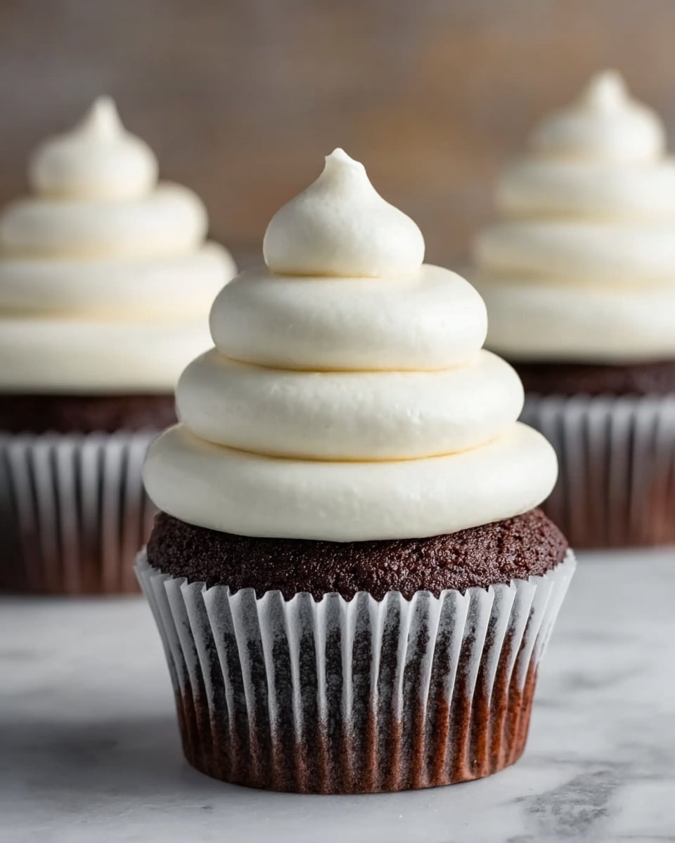 The image shows a chocolate cupcake with three thick, creamy white frosting layers stacked in a smooth, rounded cone shape on top. Each frosting layer is slightly smaller as it goes up, with the bottom one being the widest and the top ending in a soft peak. The cupcake liner is silver with a lightly crinkled texture. In the background, two more cupcakes with the same frosting design are visible but out of focus, all placed on a white marbled surface. photo taken with an iphone --ar 4:5 --v 7