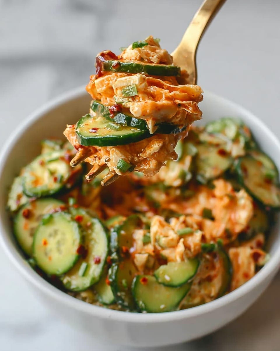 The image shows a close-up of a golden fork lifting a mix of food from a white bowl with a white marbled surface underneath. The food has layers of green cucumber slices with dark green skin and light green flesh, mixed with creamy orange pieces that have a soft texture. There are small bits of green herbs sprinkled throughout the mixture, and the food looks coated with a shiny sauce that has a reddish-brown color with hints of chili flakes. The bowl below is filled with the same mix, combining the fresh cucumber slices and creamy, orange sauce-coated pieces. Photo taken with an iphone --ar 4:5 --v 7