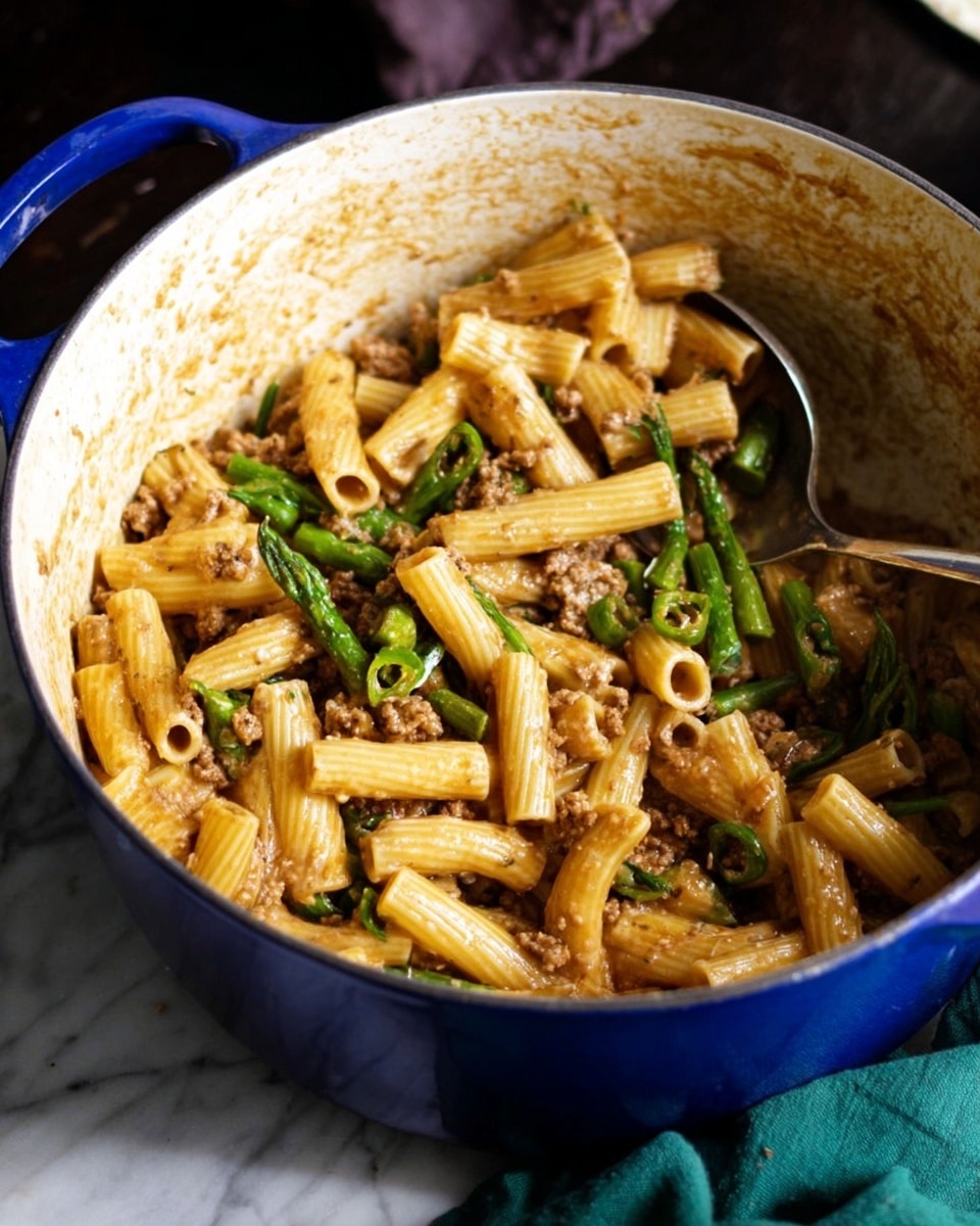A close-up view of rigatoni pasta mixed with small pieces of cooked brown meat and green vegetable stalks, all coated with light brown sauce inside a white pot with blue exterior edges. The pasta is tube-shaped with ridges and is stirred with a metal spoon partially buried in the dish, showing a mix of the ingredients evenly spread. The inside of the pot shows some cooked sauce residue around the sides, and the background features a dark surface with part of a green cloth visible. Photo taken with an iphone --ar 4:5 --v 7