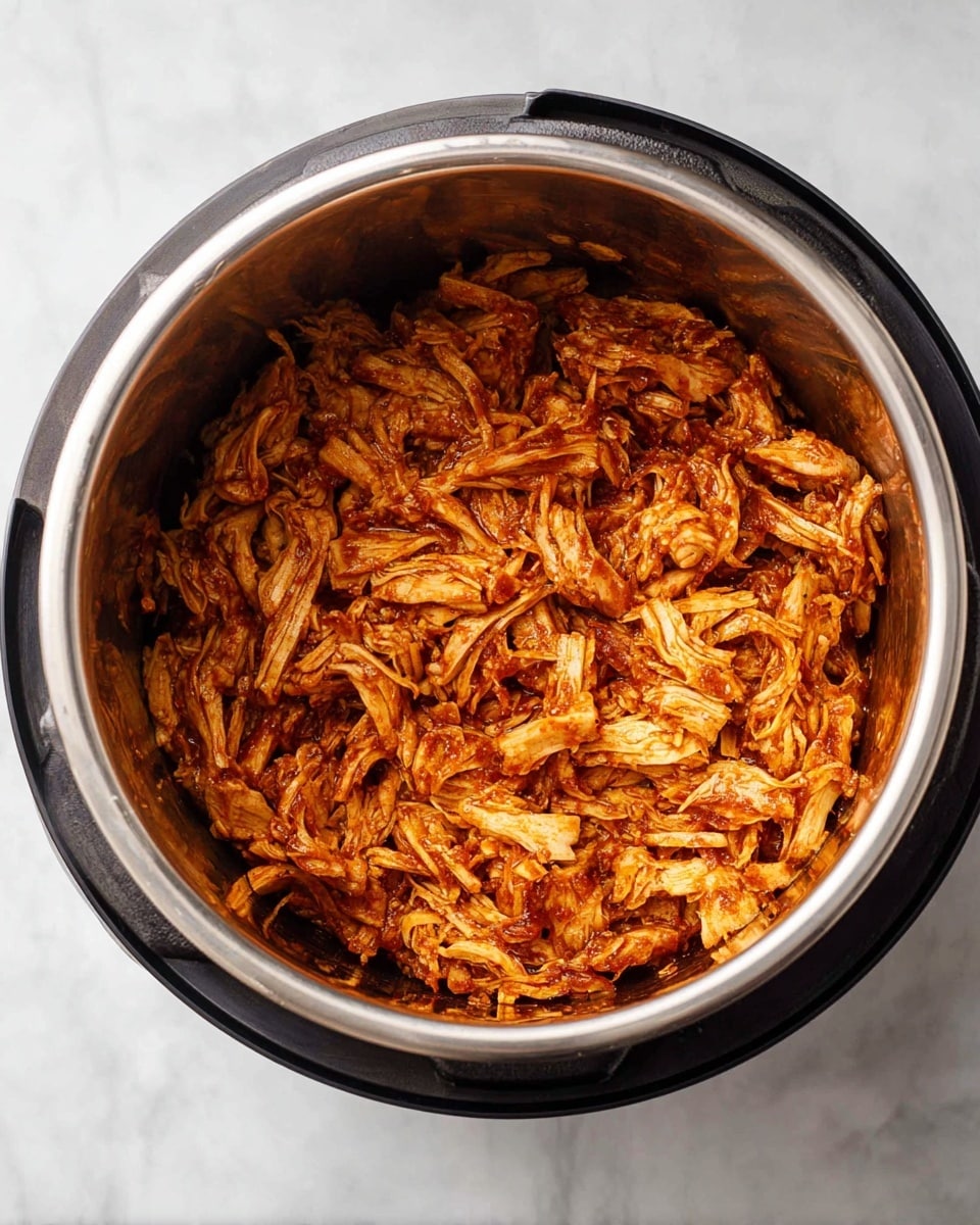 The image shows a close-up top view of a stainless steel inner pot filled with shredded cooked chicken that is evenly coated in a rich, reddish-brown sauce. The chicken pieces have a tender, fibrous texture and are mixed well, creating a dense layer that covers the entire surface inside the pot. The pot itself is part of a pressure cooker, resting on a white marbled surface, and the shiny metal of the pot reflects some light, adding a slight gloss to the scene. photo taken with an iphone --ar 4:5 --v 7