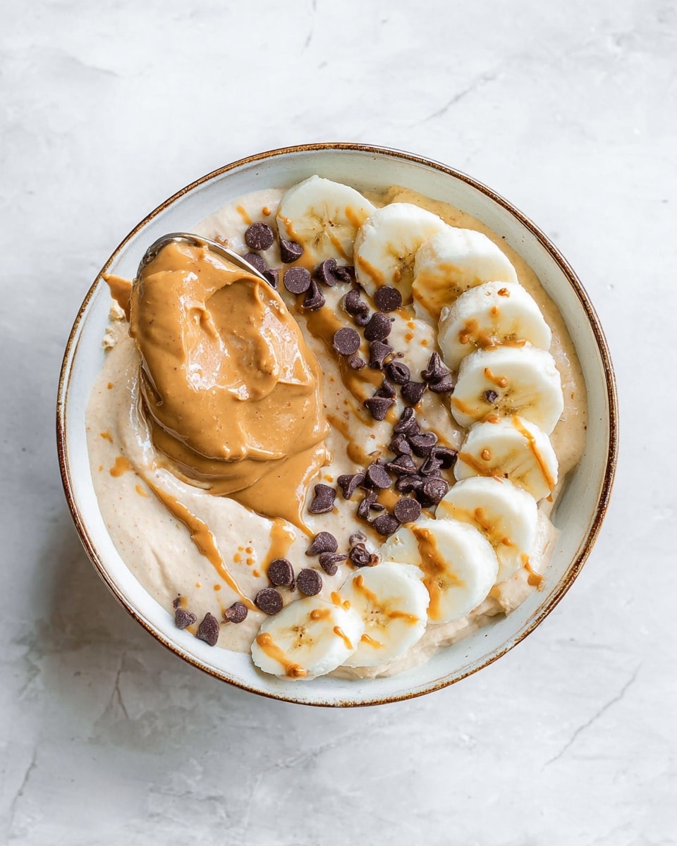 A bowl filled with a creamy beige spread as the base layer, topped with a large spoonful of smooth peanut butter placed slightly off-center. On one side of the peanut butter, there is a neat row of sliced banana pieces arranged in a curved line, some drizzled with peanut butter. Scattered over the peanut butter and bananas are small dark chocolate chips. The bowl is white with a subtle brown rim and sits on a white marbled surface. photo taken with an iphone --ar 4:5 --v 7