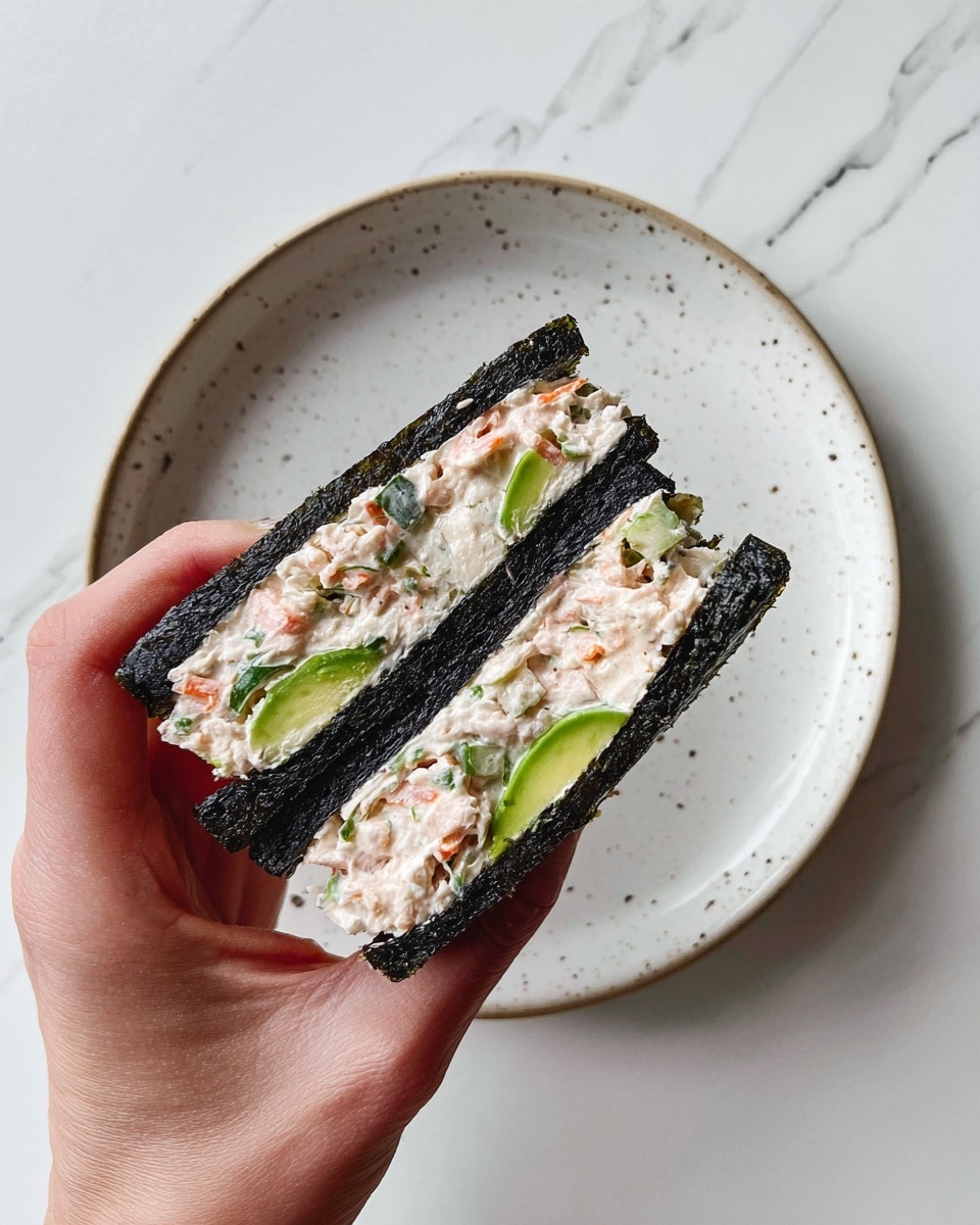 A square sheet of dark green seaweed lies flat on a white plate with small speckles. In the center of the seaweed, there is a thick layer of a creamy mixture with visible small chunks of light pink, orange, and green ingredients, giving it a textured appearance. On top of the creamy mix, four slices of bright green avocado are laid out in a neat row, slightly overlapping each other from top to bottom. The plate rests on a white marbled surface. Photo taken with an iphone --ar 4:5 --v 7