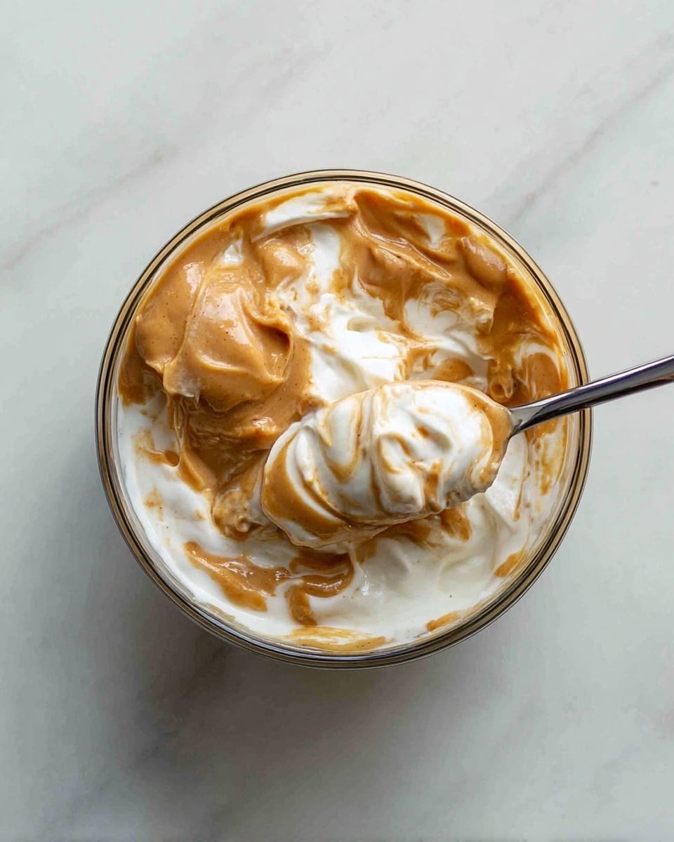 A glass bowl filled with a mix of thick white yogurt and light brown peanut butter swirled together, creating a marbled look of creamy white and rich caramel shades throughout. A metal spoon is dipped into the bowl, lifting a scoop that shows both the smooth, dense peanut butter and the airy, soft yogurt blended in swirls. The bowl is placed on a white marbled surface. Photo taken with an iphone --ar 4:5 --v 7