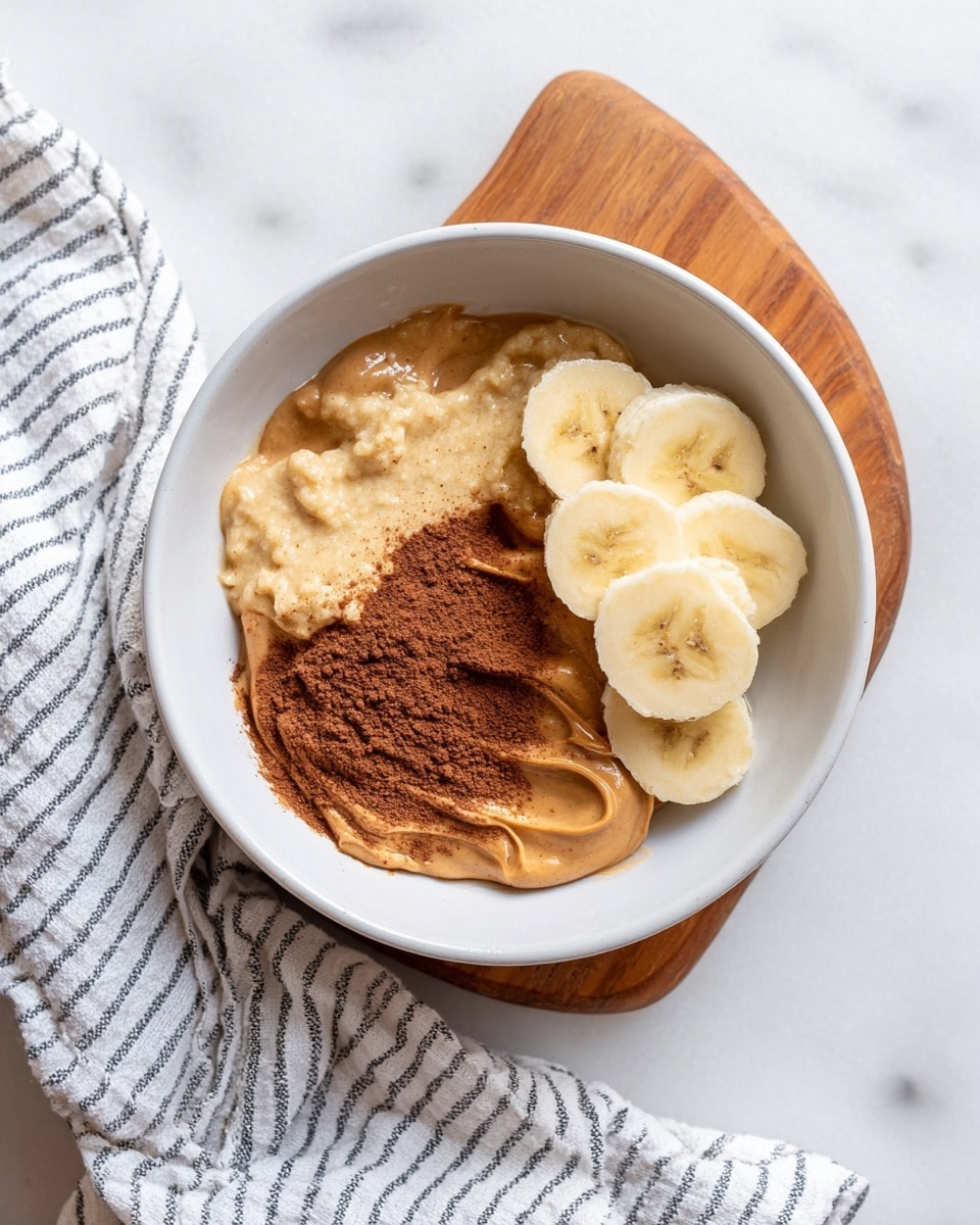 In a white bowl, mashed banana is placed on the right side with a soft, creamy texture and pale yellow color. Next to it, on the left bottom side, is a smooth layer of light brown peanut butter with a glossy surface. Above the peanut butter, a smaller amount of darker brown spread, likely a nut butter or cocoa paste, adds a thick and shiny contrast. On the left top side of the bowl, a sprinkle of brown powder, probably cinnamon, adds a fine, dusty texture. The bowl rests on a wooden board over a white marbled surface with a striped white cloth nearby. Photo taken with an iphone --ar 4:5 --v 7