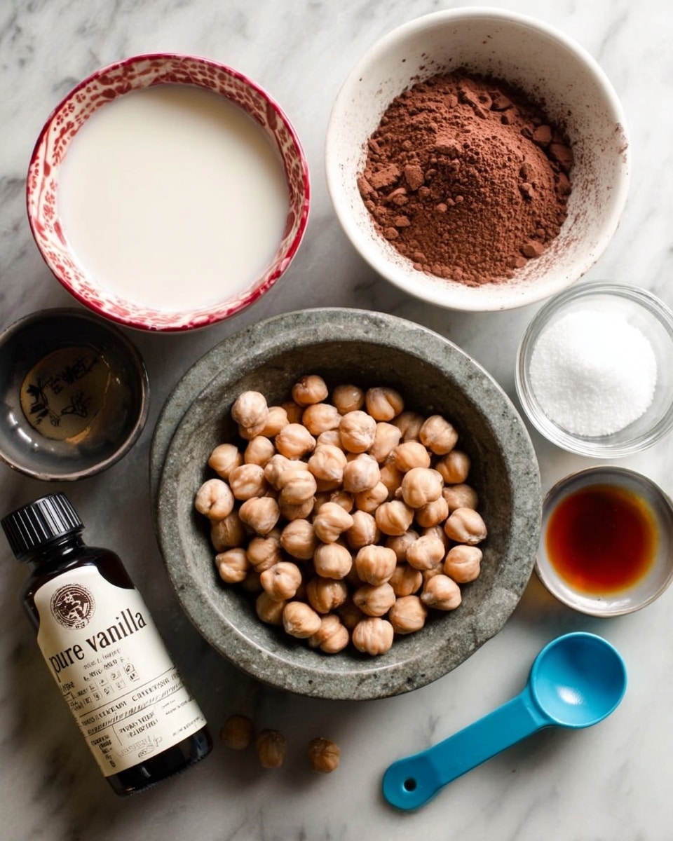 The image shows six small bowls and bottles arranged on a white marbled surface. In the center, there is a gray stone bowl filled with round, light brown hazelnuts. Above it, a white bowl is filled with dark brown cocoa powder. To the left, a white bowl with a red rim contains a white liquid, likely milk. To the right of the cocoa powder bowl, there is a smaller white bowl holding a dark amber liquid, probably vanilla extract. Above the milk bowl, there is a tiny clear bowl containing white granulated sugar. In the bottom left corner, a white and black bottle labeled