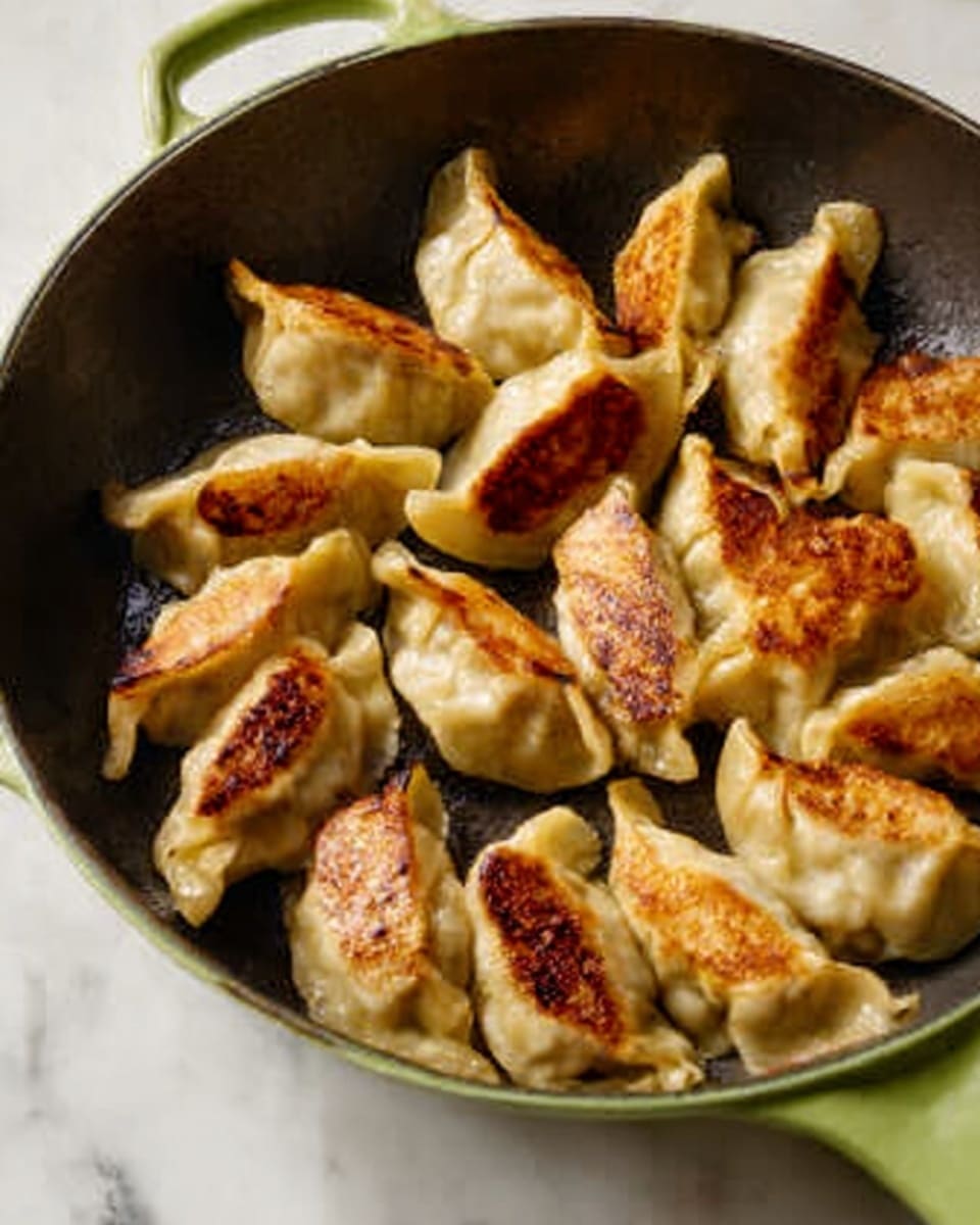 The image shows a green cast iron pan filled with one layer of golden-brown dumplings. Each dumpling has a crispy, browned side facing up, with the rest of the dough a light beige color and slightly crinkled edges. The pan sits on a white marbled surface, and the dumplings are arranged loosely but close to each other, taking up most of the pan’s bottom. Photo taken with an iphone --ar 4:5 --v 7