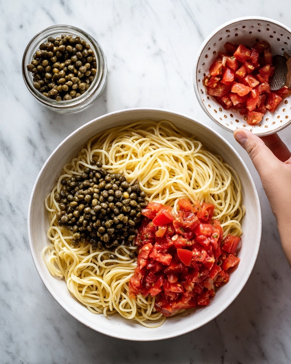 A white bowl filled with three layers of food sits on a white marbled surface. The bottom layer is light yellow cooked spaghetti noodles, thin and tangled. On the right side of the noodles is a layer of small red diced tomatoes, fresh and slightly shiny. On the left side is a layer of dark green capers, small and round, adding texture contrast. Above the bowl, a woman's hand holds a small white bowl with a dotted pattern, pouring more diced tomatoes onto the top right of the noodles. Nearby is an open jar of capers. The photo taken with an iphone --ar 4:5 --v 7