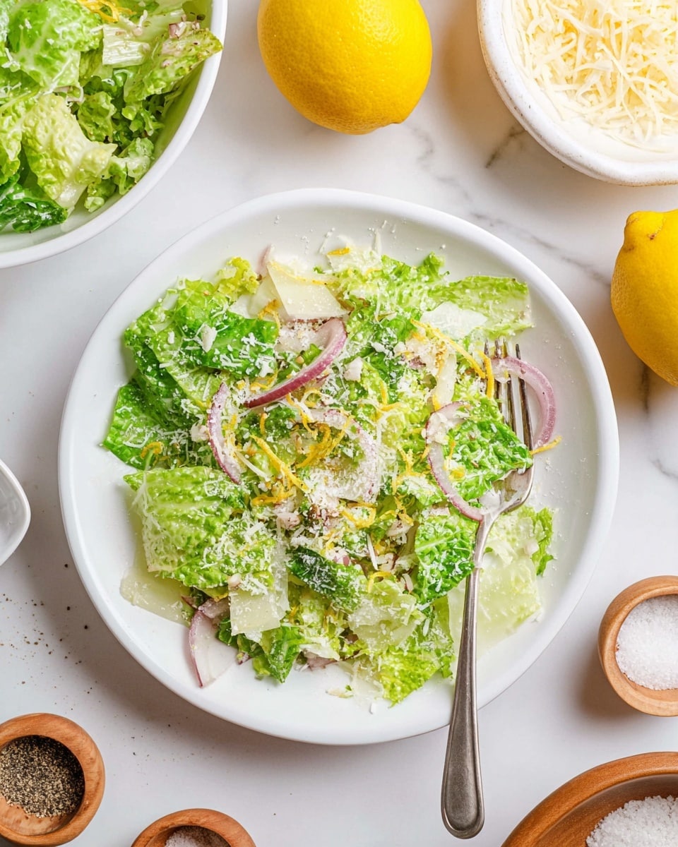 A white round plate holds a light salad with two main layers: the bottom layer is made of chopped green lettuce with various shades from light to medium green, and the top layer is sprinkled with thin slices of red onion, fine shreds of pale yellow cheese, and thin yellow lemon zest strands, giving texture and contrast. A silver fork rests on the right side of the plate, partially inside the salad. The plate sits on a white marbled surface, surrounded by two whole bright yellow lemons at the top right and bottom left, a larger white bowl filled with more lettuce salad at the top, a white dish with shredded cheese at the top right, and two small wooden spoons holding coarse salt and ground black pepper at the bottom center. Photo taken with an iphone --ar 4:5 --v 7