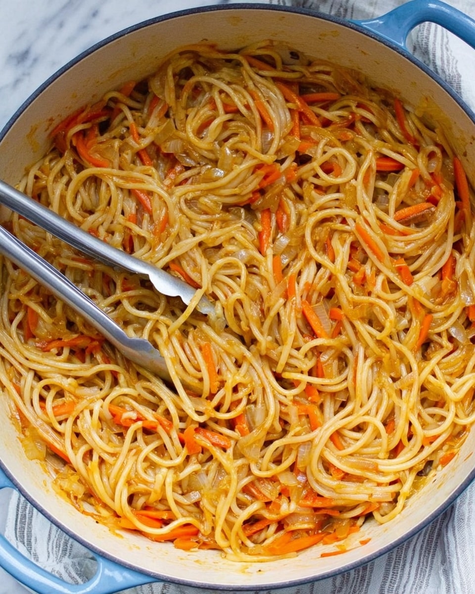 A close-up view of a pot filled with spaghetti noodles mixed evenly with thin strips of orange carrots and small pieces of translucent onions, all coated in a light orange sauce. The noodles look soft and slightly shiny, while a pair of silver tongs rests on the left side inside the pot, ready to serve. The pot is white with light blue on the outside, sitting on a white marbled surface with a light striped cloth underneath. The overall look is warm and inviting, showing the texture of the noodles and vegetables clearly, photo taken with an iphone --ar 4:5 --v 7