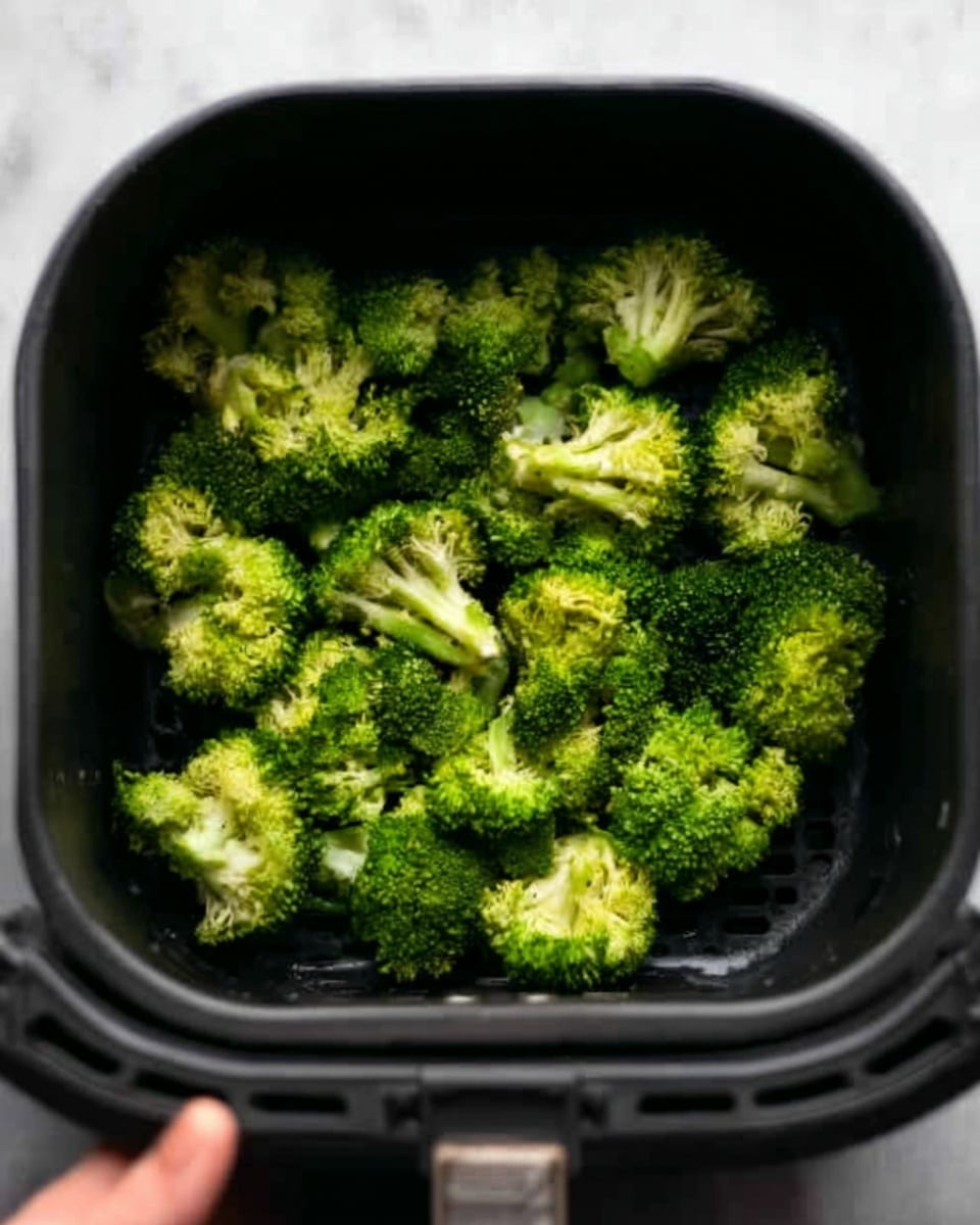A black air fryer basket filled with bright green broccoli florets, each floret showing a dense, bumpy texture and slightly varying in size, arranged closely together inside the basket. The basket has small round holes at the bottom for air circulation. The image shows the broccoli from above, with a woman’s hand holding the basket handle on the edge. The background is a white marbled texture. Photo taken with an iphone --ar 4:5 --v 7