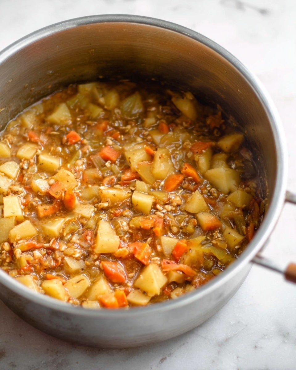 The image shows a silver pot filled with a thick stew that has three main visible layers. The top layer is made up of small chunks of light yellow and orange vegetables, likely potatoes and carrots, mixed with bits of beige-colored meat or grains throughout. The middle layer is a light brown, slightly shiny liquid that coats all the ingredients, giving the stew a wet and hearty look. The bottom layer is mostly hidden but appears to have more chunks and liquid blending together. The pot is placed on a white marbled surface with soft, natural lighting highlighting the textures inside the pot. photo taken with an iphone --ar 4:5 --v 7
