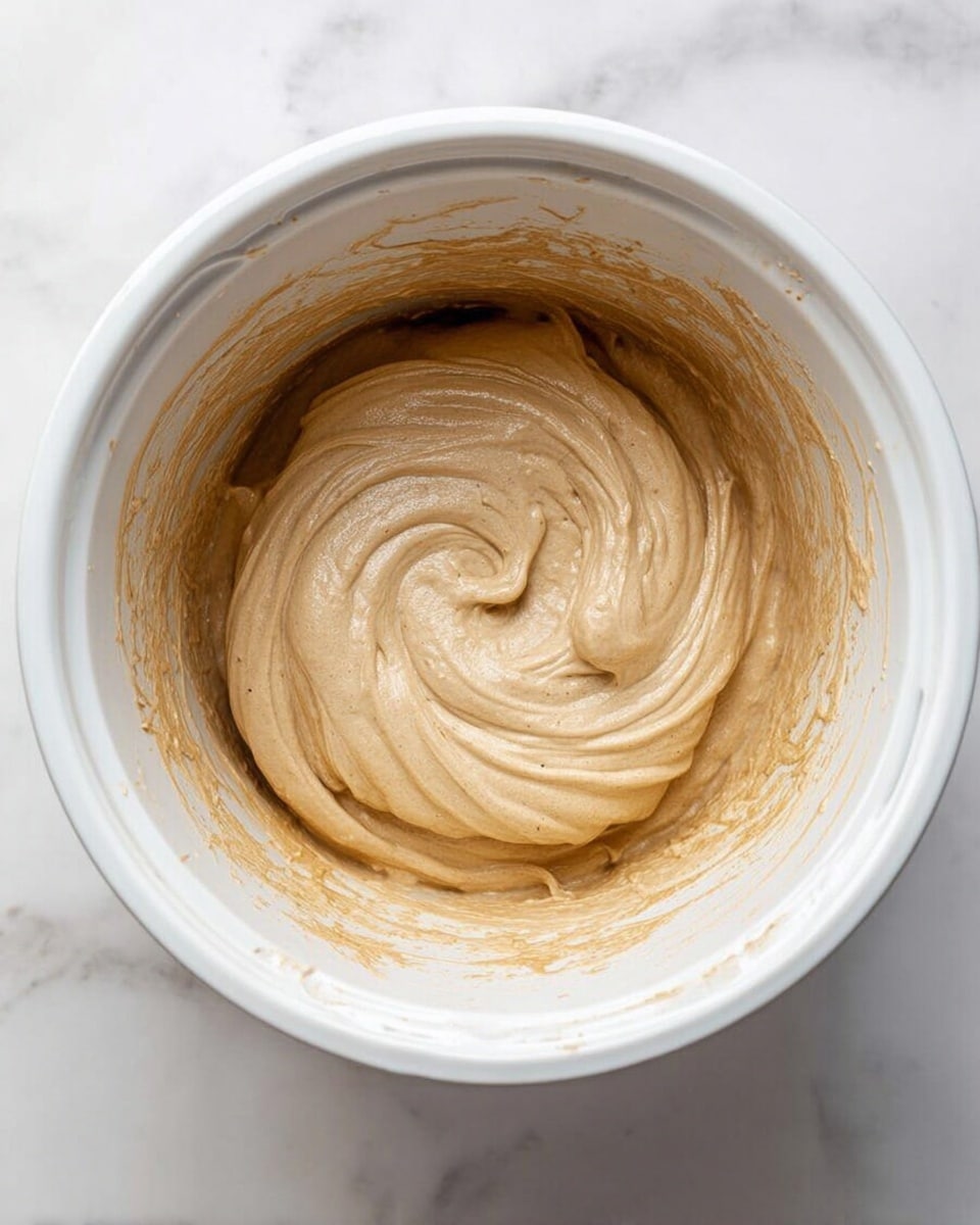 A close-up view of a creamy light brown ice cream with small dark specks mixed in, shown inside a white rectangular container. A metal ice cream scoop is lifting a round, smooth scoop of the ice cream from the container, creating soft, textured waves on the surface. The container is placed on a white marbled surface, and a woman's hand is holding the scoop. In the background, there is a small white cup with a dark liquid inside, slightly out of focus. photo taken with an iphone --ar 4:5 --v 7