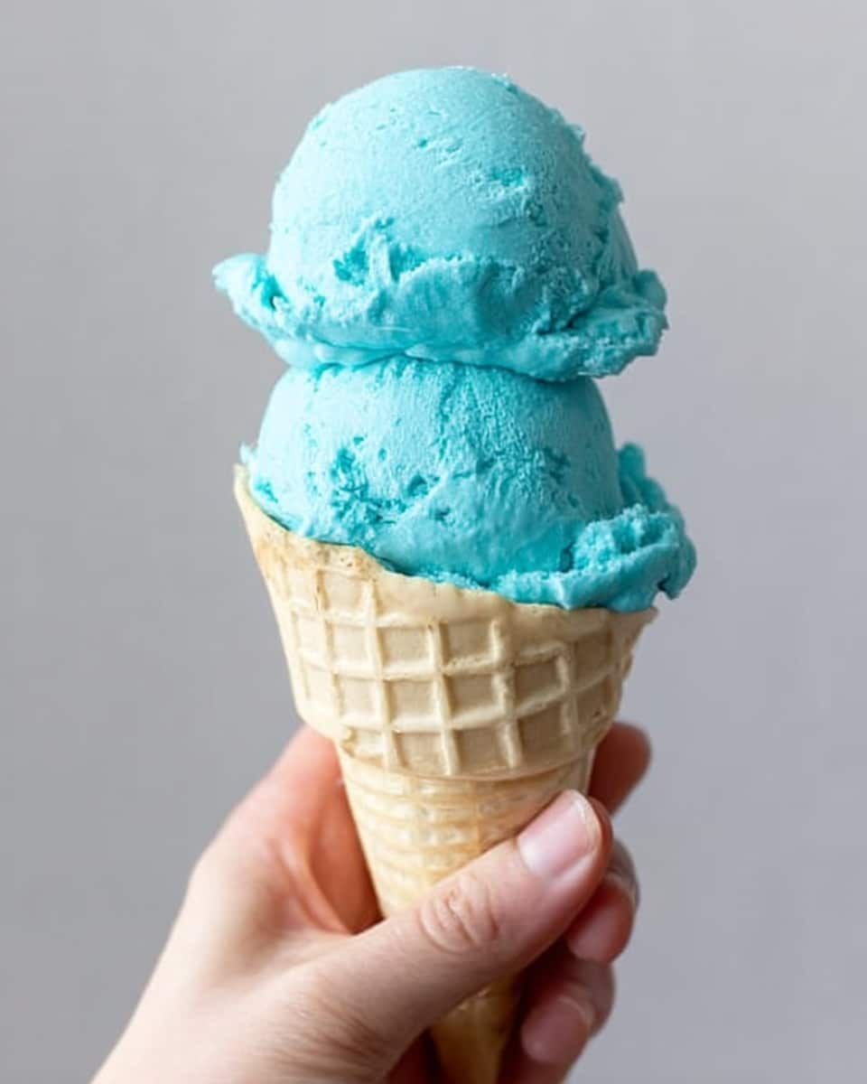 A woman's hand is holding a waffle cone filled with two large scoops of bright blue ice cream stacked on top of each other. The ice cream has a smooth and creamy texture with some small ridges and swirls showing its softness. The cone is light beige with a grid pattern, and the background is a plain light gray, making the blue ice cream stand out clearly. photo taken with an iphone --ar 4:5 --v 7