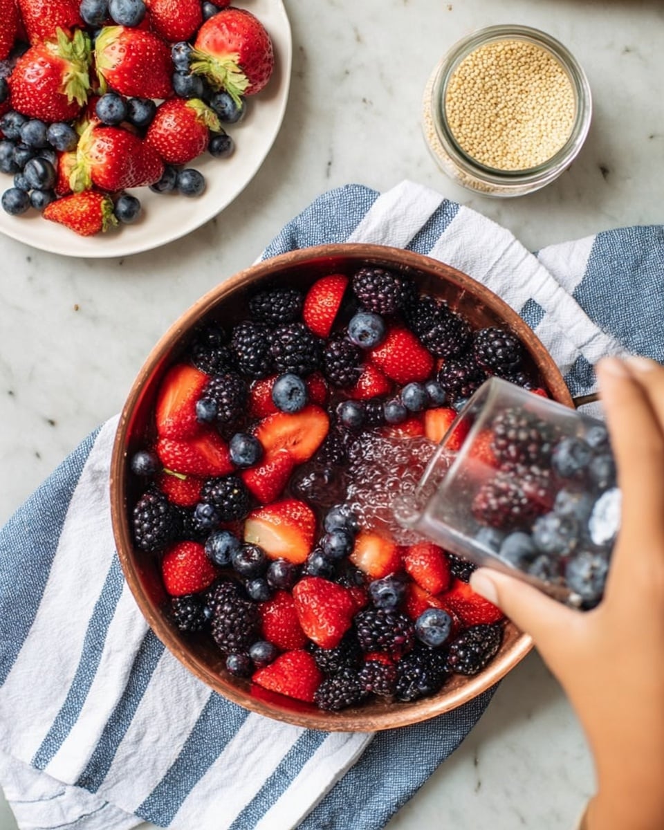 A round copper pan filled with bright red strawberries, deep blue blueberries, and black blackberries, all mixed together showing vibrant colors and glossy textures. The pan is on a white and blue striped cloth lying on a white marbled surface. A woman's hand is holding a clear glass container, pouring more berries into the pan. At the top left corner, a white plate is filled with whole strawberries, blueberries, and blackberries, with the fruit looking fresh and ripe. A small jar with beige seeds is placed on the top right on the white marbled surface. Photo taken with an iphone --ar 4:5 --v 7