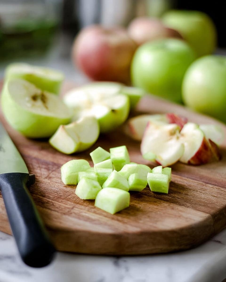 The image shows a wooden cutting board with pieces of peeled green apple on it. In the front center, there are small cubes of green apple neatly grouped together. Behind the cubes, there are larger peeled apple sections, some with a bit of red skin left on the edges. To the left, there is a black-handled knife laying flat on the board. In the soft background, there are whole apples out of focus, giving a warm kitchen feel. The whole scene is set on a white marbled surface. photo taken with an iphone --ar 4:5 --v 7