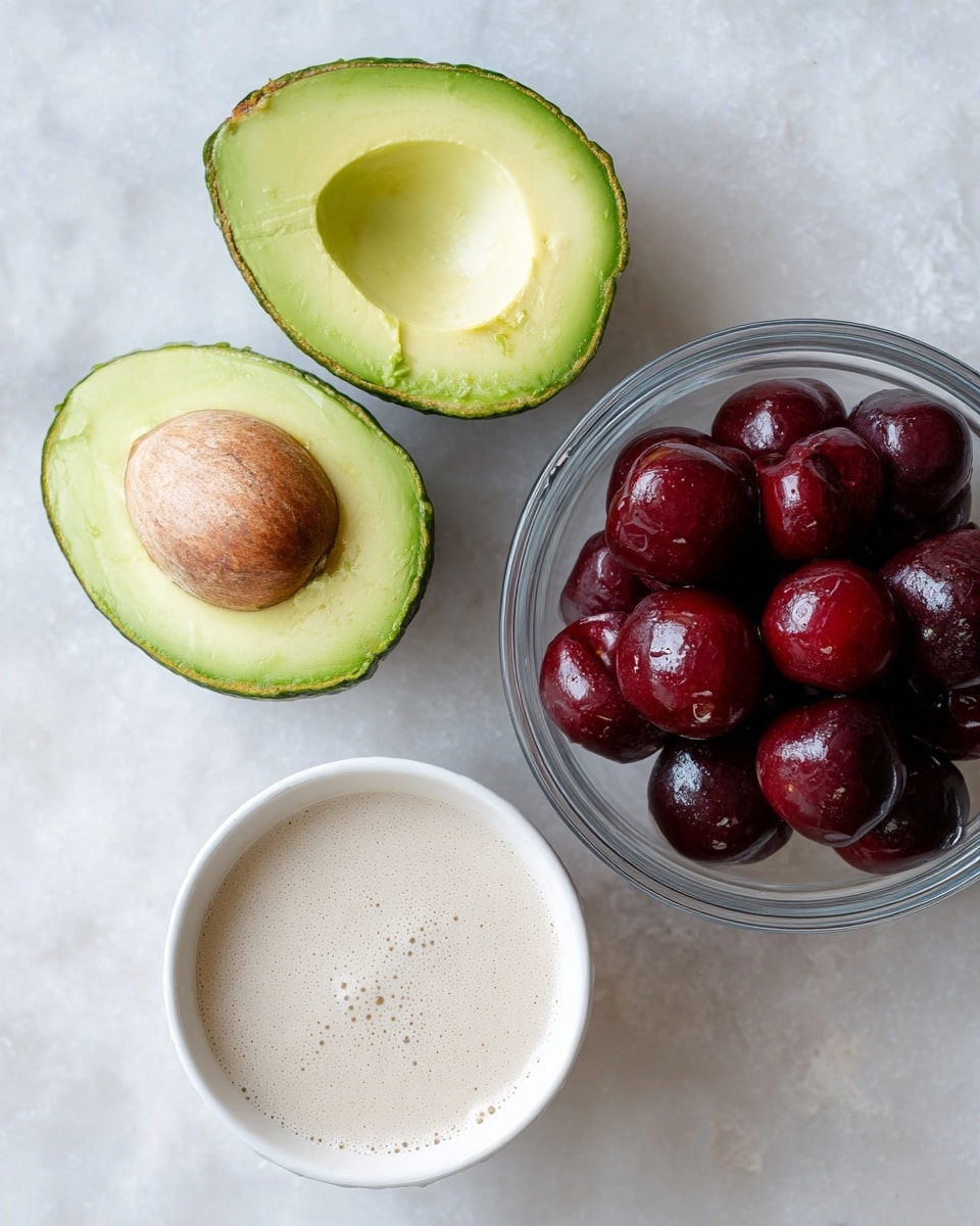 The image shows three main food items placed on a white marbled surface. On the left side, there is a halved avocado with one half showing the brown seed in the center and the other half showing the smooth light green inside flesh. To the right is a white bowl filled with a creamy, pale beige liquid with small bubbles on the surface. Below and slightly to the right, there is a glass bowl filled with dark red cherries, some of which appear shiny and fresh. The composition is simple and clean, with each item clearly visible and distinct from the white marbled background. photo taken with an iphone --ar 4:5 --v 7