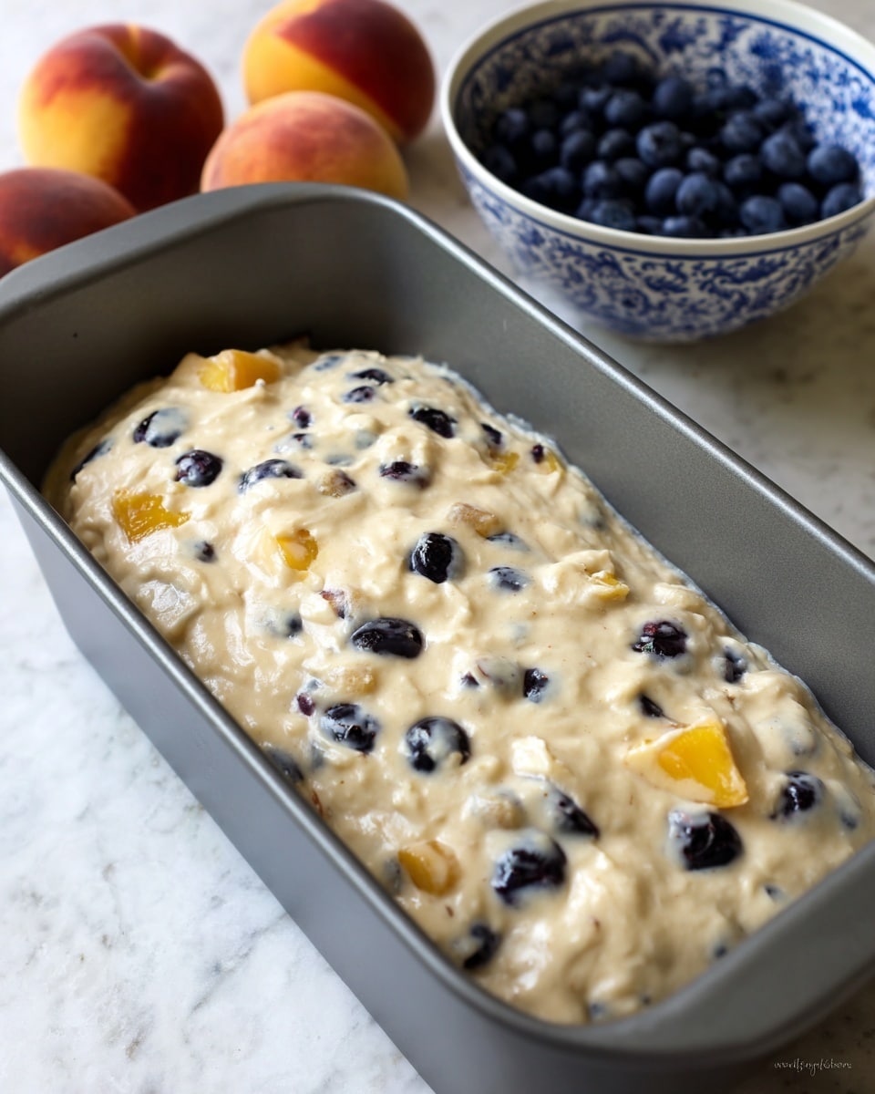 A gray loaf pan filled with a thick, creamy batter mixed with blueberries and chunks of yellow fruit, likely peaches. The batter is light beige with soft textures, and the blueberries create scattered dark blue spots across the surface. In the background, four peaches rest next to a white bowl with a blue intricate pattern filled with more blueberries, all placed on a white marbled surface. Photo taken with an iphone --ar 4:5 --v 7