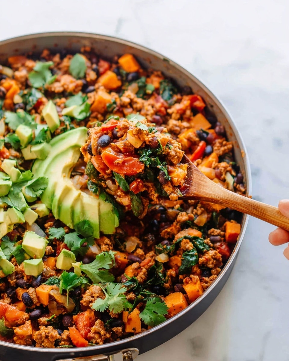 The image shows a close-up of a pan filled with a colorful mix of cooked ingredients. The main layer is a combination of small orange cubes, black beans, leafy green spinach, and chunks of browned ground meat mixed with diced red tomatoes. On top of this, there are small pieces of bright green avocado and fresh cilantro leaves scattered around. A woman's hand is holding a wooden spoon lifting a spoonful of the mixture, showing the texture of the cooked ingredients clearly. The pan is placed on a white marbled surface that adds a clean, fresh look to the image. photo taken with an iphone --ar 4:5 --v 7