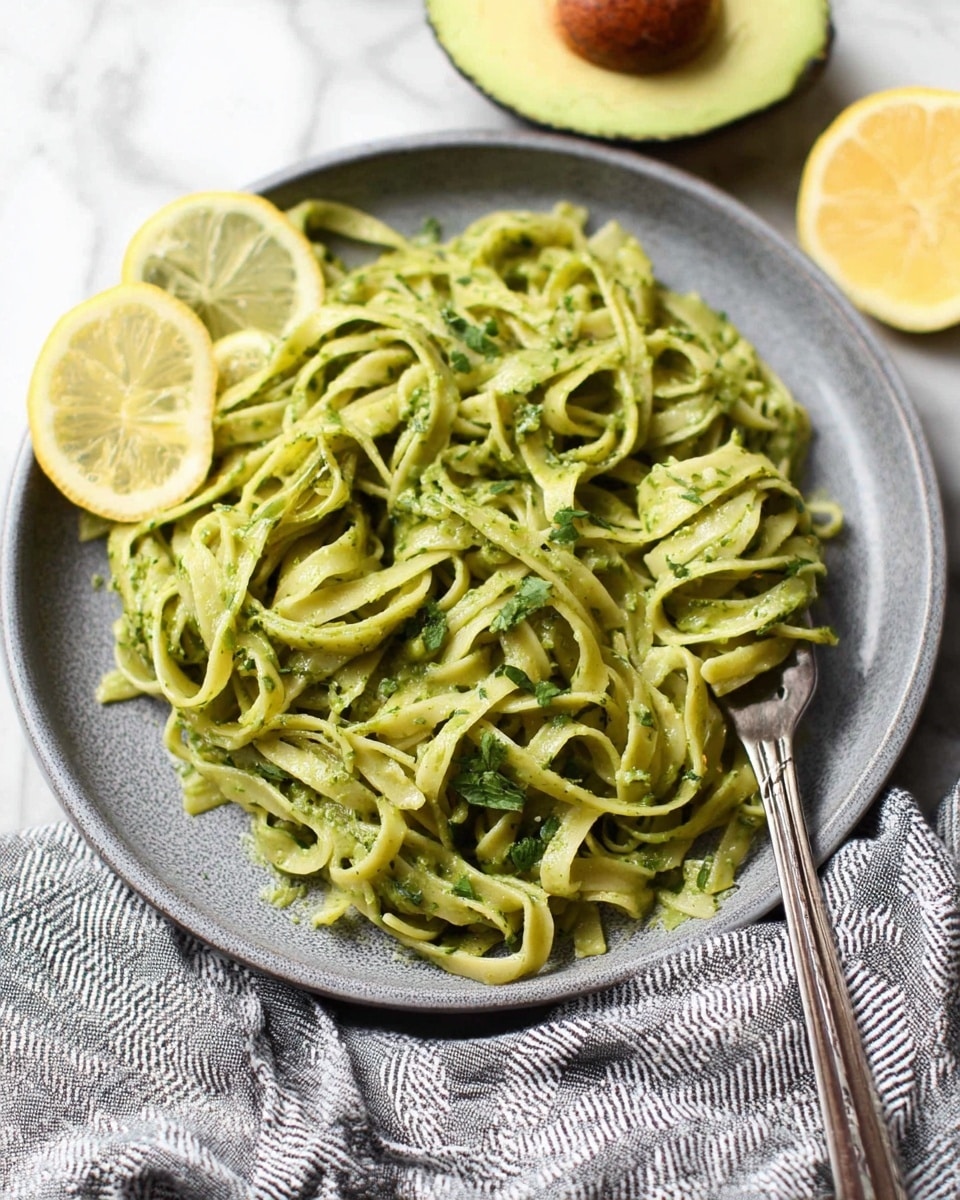 The image shows a close-up of wide pasta ribbons in a creamy green sauce, likely made with avocado or spinach, giving it a smooth and rich texture. The pasta is mixed with torn dark green basil leaves, scattered throughout the dish, and a few small red chili flakes adding specks of color. A silver fork is lifting some pasta from the bowl, showing the thick sauce coating each ribbon. The pasta and sauce are placed inside a white bowl on a white marbled surface. The photo taken with an iphone --ar 4:5 --v 7