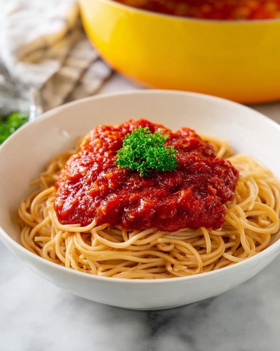A white bowl filled with a nest of plain light brown spaghetti noodles at the bottom, topped with a thick layer of chunky, bright red tomato sauce that looks warm and fresh. On top of the sauce, there is a small bright green parsley garnish adding a pop of color. The bowl sits on a white marbled surface, with a yellow pot blurred in the background. Photo taken with an iphone --ar 4:5 --v 7