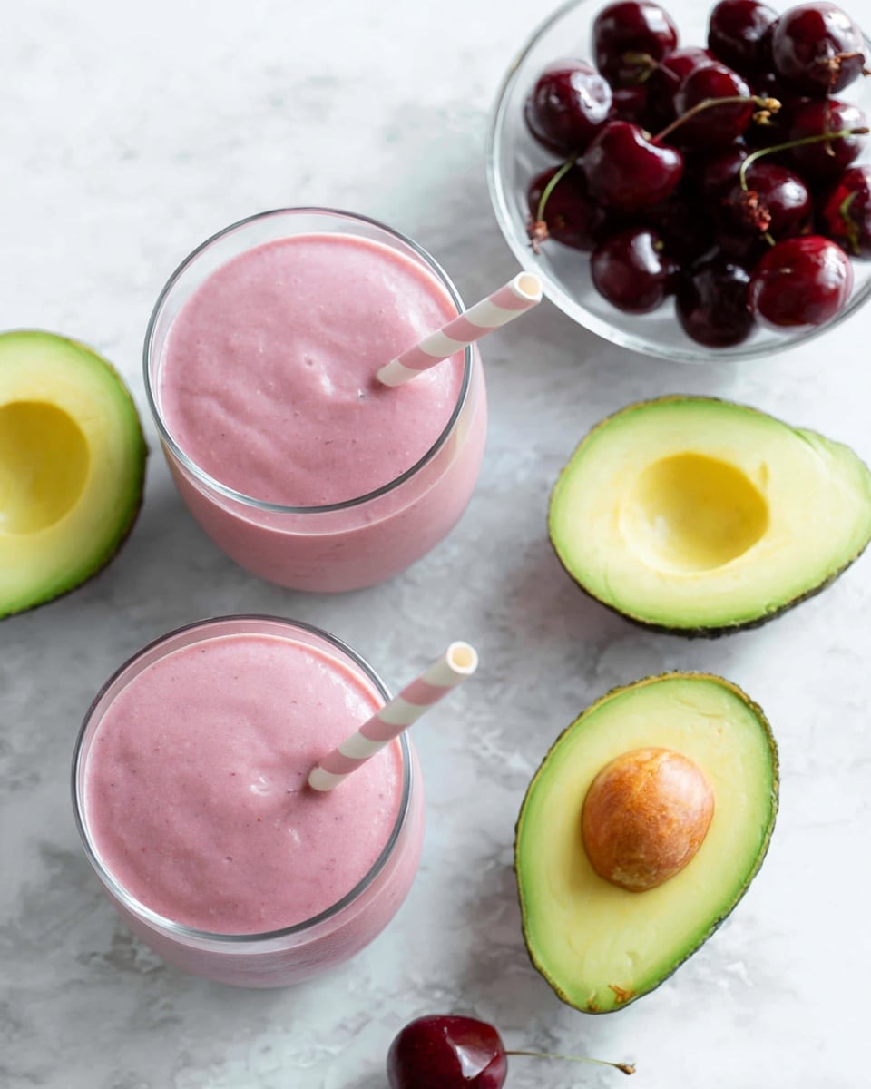The image shows two glasses filled with a smooth pink smoothie, each topped with a white and light pink striped straw. Around the glasses, there are two avocado halves: one with the pit and another without, both showing bright green and yellow colors with soft textures. A small clear glass bowl filled with dark red cherries is placed near the top right, shining under the light with a slightly wet look. All items are set on a white marbled surface that brightens the scene with a soft texture. Photo taken with an iphone --ar 4:5 --v 7