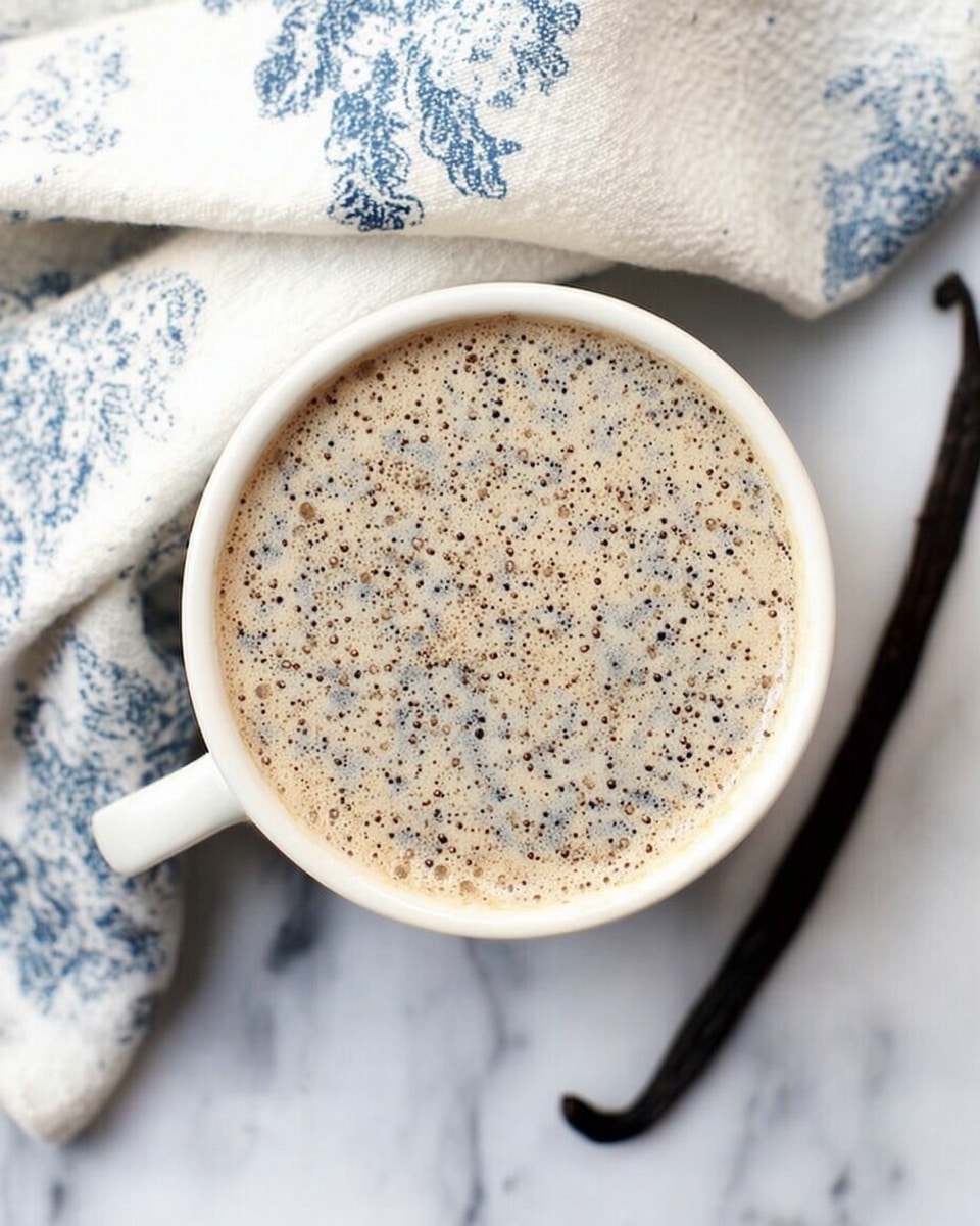 A white cup filled with a creamy light beige drink speckled with many tiny black dots on the surface, giving a textured look. The cup is placed on a white marbled surface next to a light cloth with blue patterns and a dark vanilla bean pod lying flat on the right side. Photo taken with an iphone --ar 4:5 --v 7