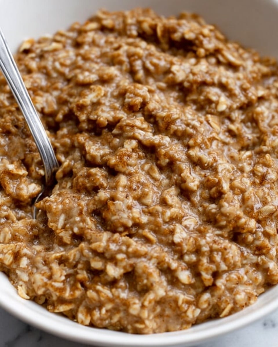 A close-up view of a bowl filled with thick oatmeal that looks creamy and brown in color, with visible oats mixed evenly throughout. A silver spoon is partially dipped into the oatmeal on the left side of the bowl. The bowl is white and sits on a white marbled surface. The texture of the oatmeal is slightly rough but soft, showing individual oats and a smooth brown mixture photo taken with an iphone --ar 4:5 --v 7