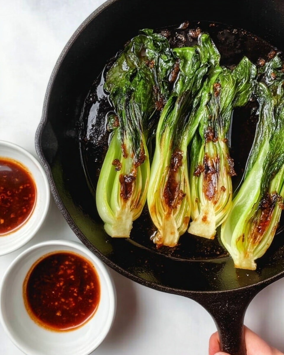 The image shows four cooked baby bok choy lying in a dark cast iron pan, each with bright green leaves and pale green stalks, slightly browned on the sides from cooking. Next to the pan on the white marbled surface are two small white bowls, each filled with a dark reddish-brown sauce. A woman's hand is holding the pan by its handle. The photo taken with an iphone --ar 4:5 --v 7