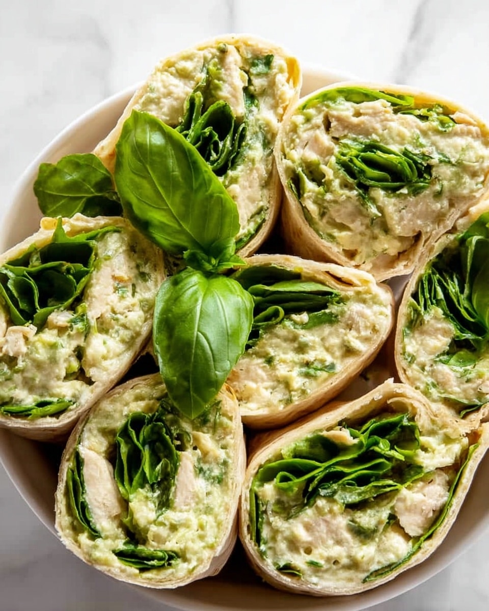 The image shows a close-up of several tortilla wraps cut into halves and arranged neatly in a white bowl. Each wrap has three visible layers: the outer soft, light beige tortilla, a middle layer of fresh, bright green spinach leaves, and an inner creamy light green spread mixed with small pieces of white chicken. A few fresh basil leaves are placed on top for garnish. The bowl is set on a white marbled surface. Photo taken with an iphone --ar 4:5 --v 7