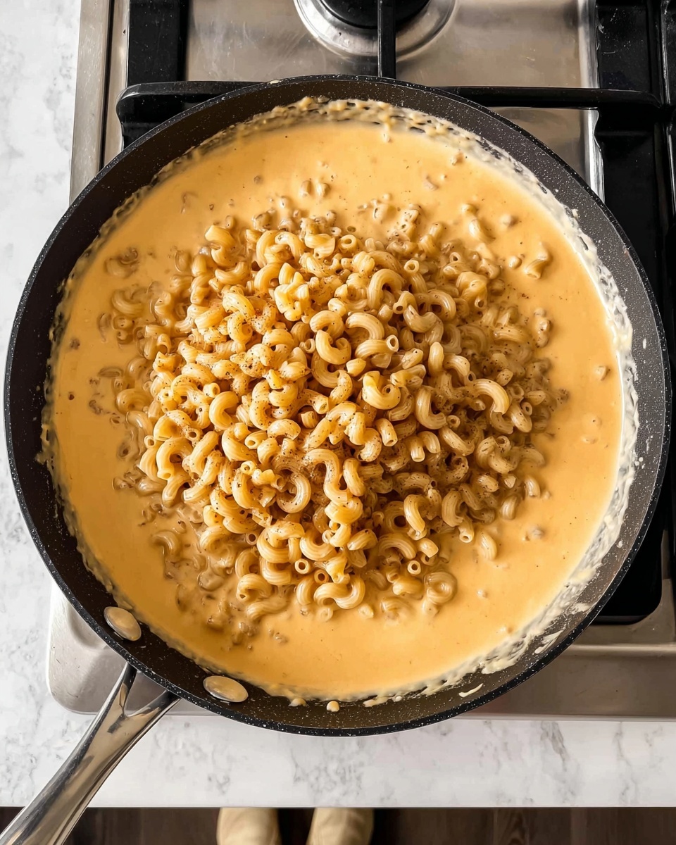 The image shows a black pan on a stove with cooked elbow pasta piled in the center, surrounded by creamy, light orange cheese sauce that coats the bottom and sides of the pan. The pasta forms a mound with small curved tubes, contrasting with the smooth and thick sauce beneath it. The stove is silver and black, and the surface around it is a white marbled texture, with a hint of someone's beige shoes visible at the bottom edge. Photo taken with an iphone --ar 4:5 --v 7