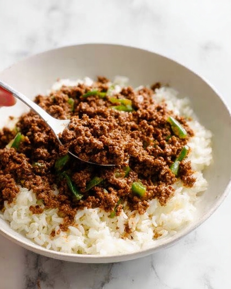 A white bowl filled with a base layer of fluffy white rice, with a thick layer of cooked ground beef topped on it. The beef is brown and has some visible green vegetable pieces mixed in, adding some color contrast. A woman's hand holding a white spoon is scooping some rice and beef from the bowl. The bowl is on a white marbled surface. photo taken with an iphone --ar 4:5 --v 7
