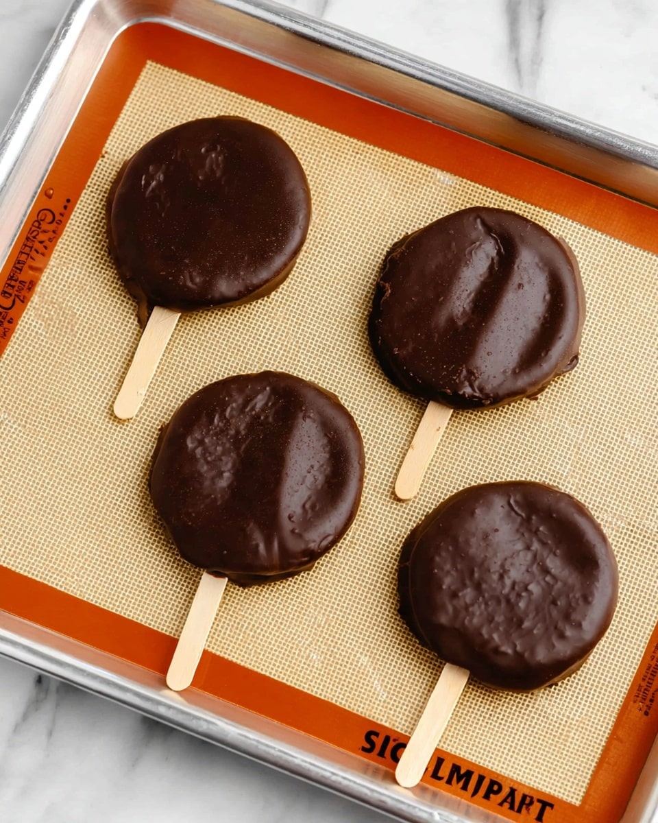 The image shows four round treats on wooden sticks, each fully covered in smooth, dark chocolate. The treats are placed on a light beige silicone baking mat with an orange border inside a silver metal baking tray. The chocolate coating has a slightly bumpy texture with a few small indentations, and the wooden sticks are light tan and extend from the bottom of each chocolate circle. The tray is resting on a white marbled surface. photo taken with an iphone --ar 4:5 --v 7