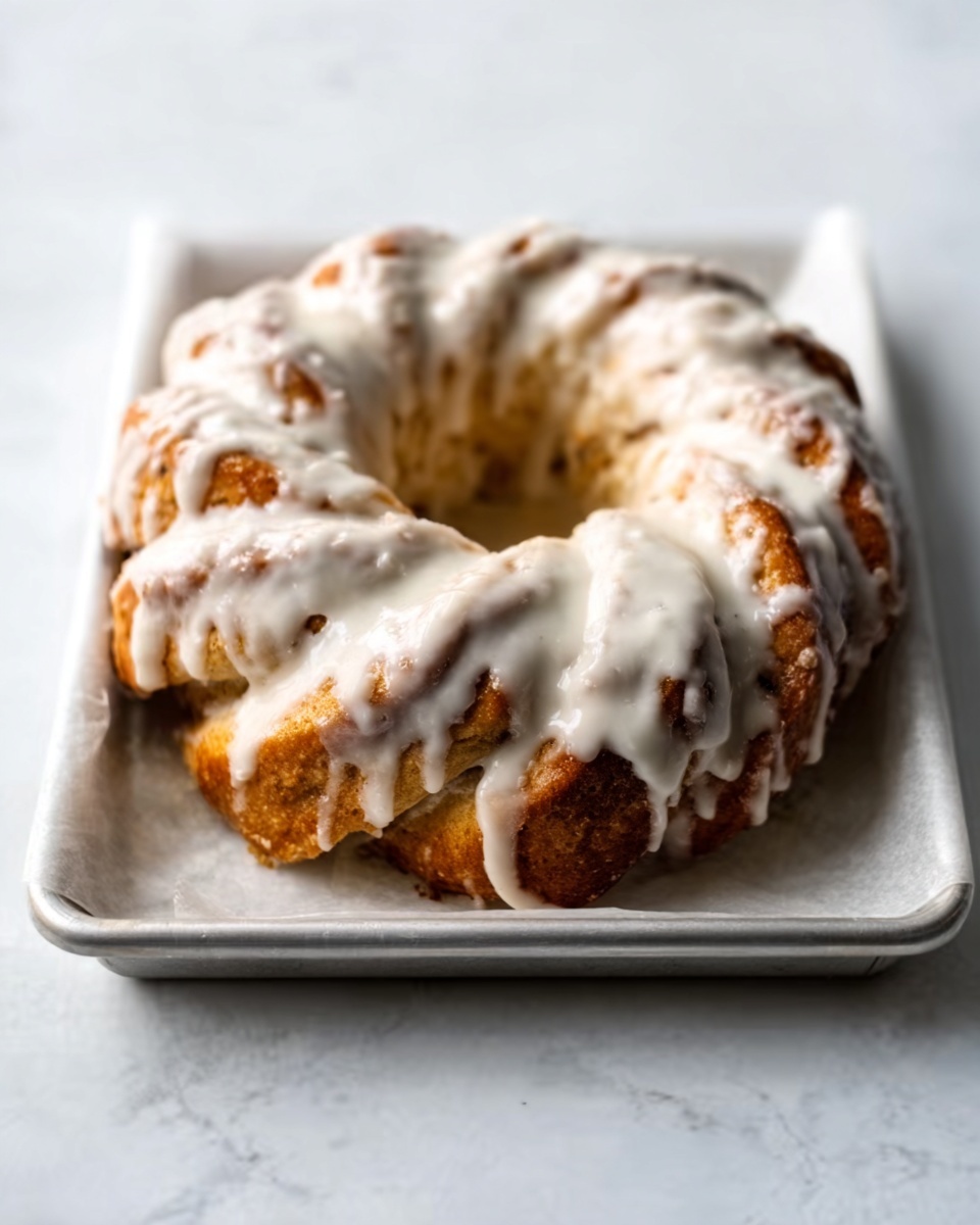 A twisted bread ring sits on a white tray covered with white parchment paper. The bread is brown with small darker spots showing where it is baked, and it is covered with a thick layer of white icing that drips slightly down the sides. The bread’s twisted shape forms a circular pattern with a hole in the center. The background is a white marbled surface. photo taken with an iphone --ar 4:5 --v 7