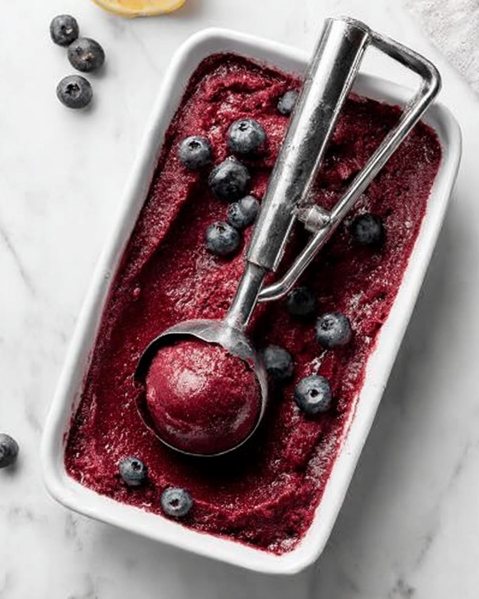 The image shows a white rectangular dish filled with deep red-purple berry sorbet, with a smooth and slightly icy texture. Several whole blueberries are scattered on top, adding small round shapes in dark blue color. There is a shiny silver ice cream scoop partly submerged in the sorbet, holding a round scoop in the center. The dish sits on a white marbled surface with a few blueberries nearby. photo taken with an iphone --ar 4:5 --v 7