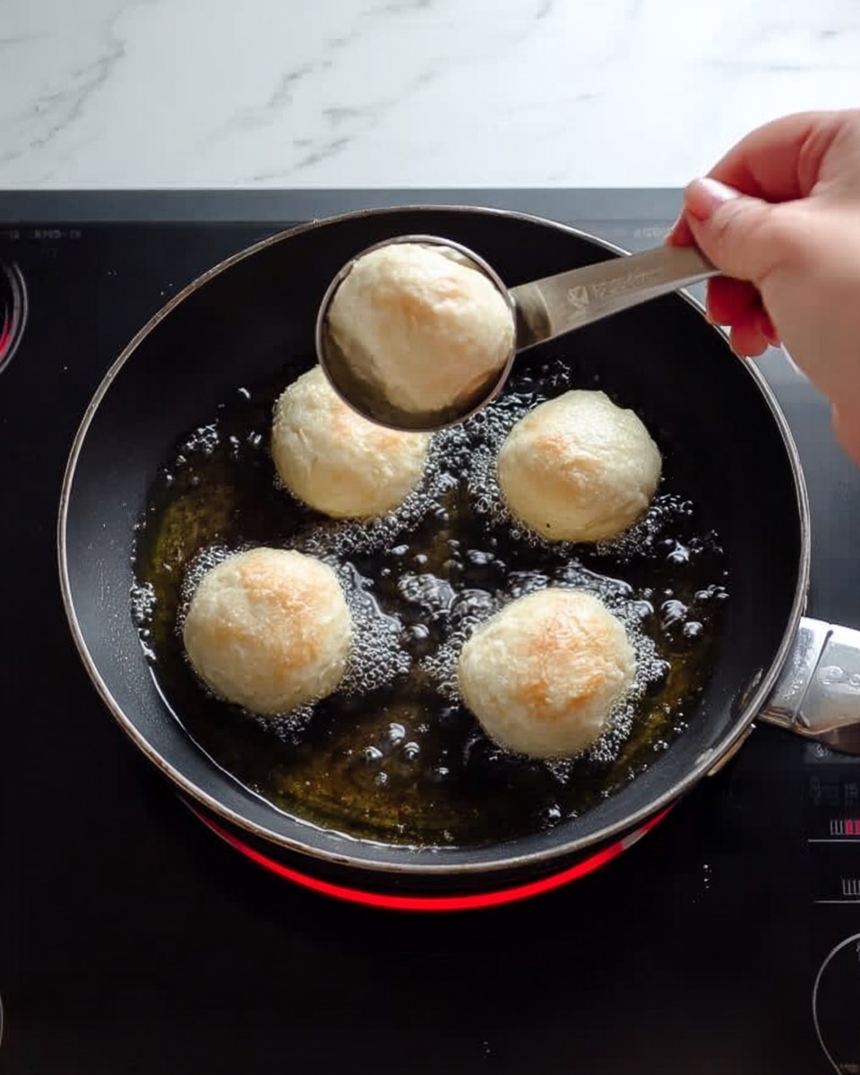 A woman's hand is holding a silver scoop filled with pale, soft dough, placing round dough balls into dark oil inside a black pan. There are three dough balls frying, light golden at the edges with small bubbles forming around them. The pan sits on a stovetop burner glowing red with heat. The background is a white marbled texture. photo taken with an iphone --ar 4:5 --v 7