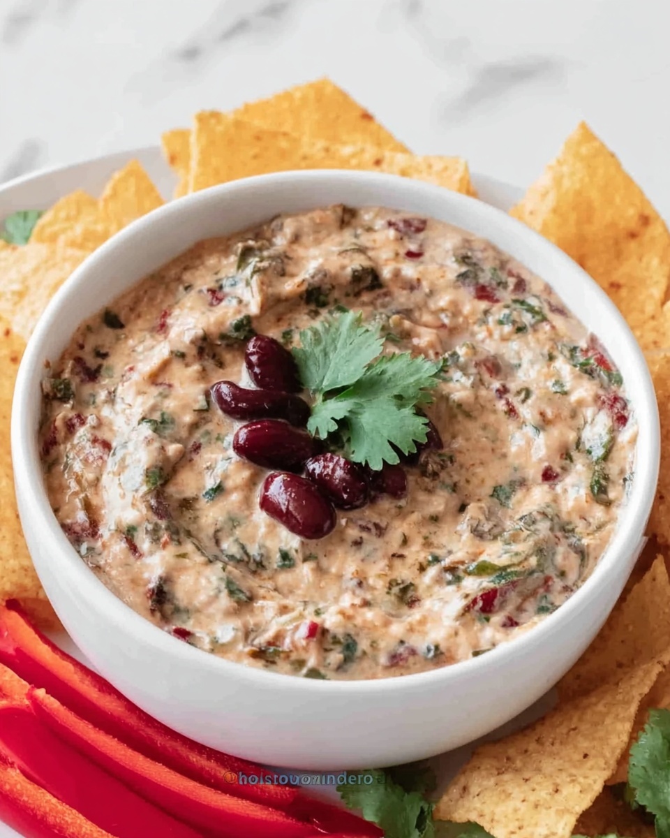 A white bowl filled with a creamy mixture that has a light beige color with visible green herbs mixed throughout. On top of the creamy layer, there are a few dark red beans and a small sprig of green cilantro placed in the center. The bowl sits on a white marbled surface surrounded by bright red bell pepper strips and light-colored tortilla chips, arranged loosely around the bowl. photo taken with an iphone --ar 4:5 --v 7